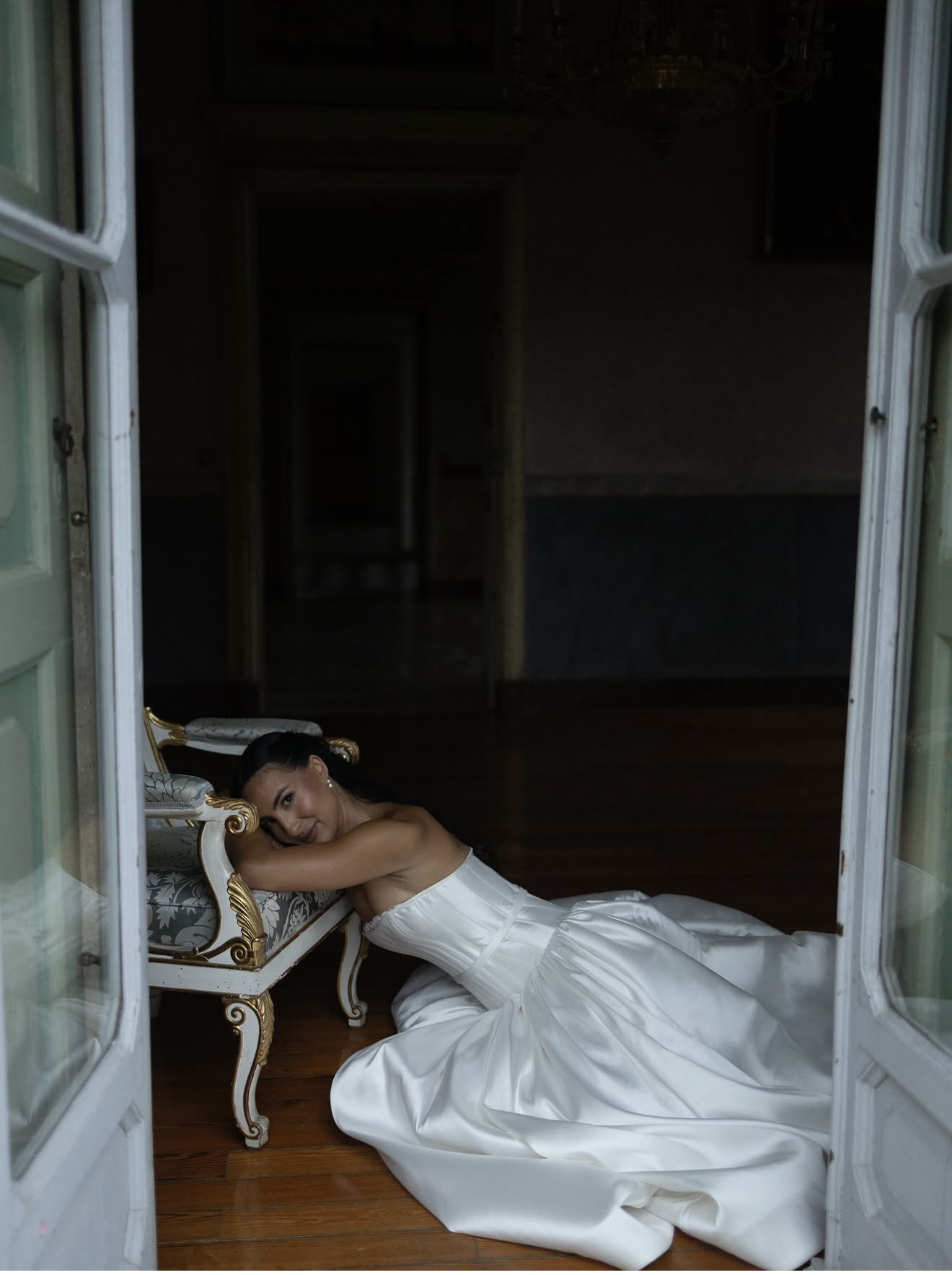 Woman in white wedding dress lying on the floor, resting her head on a decorative armchair, inside a room viewed through open double doors in Lake Como.