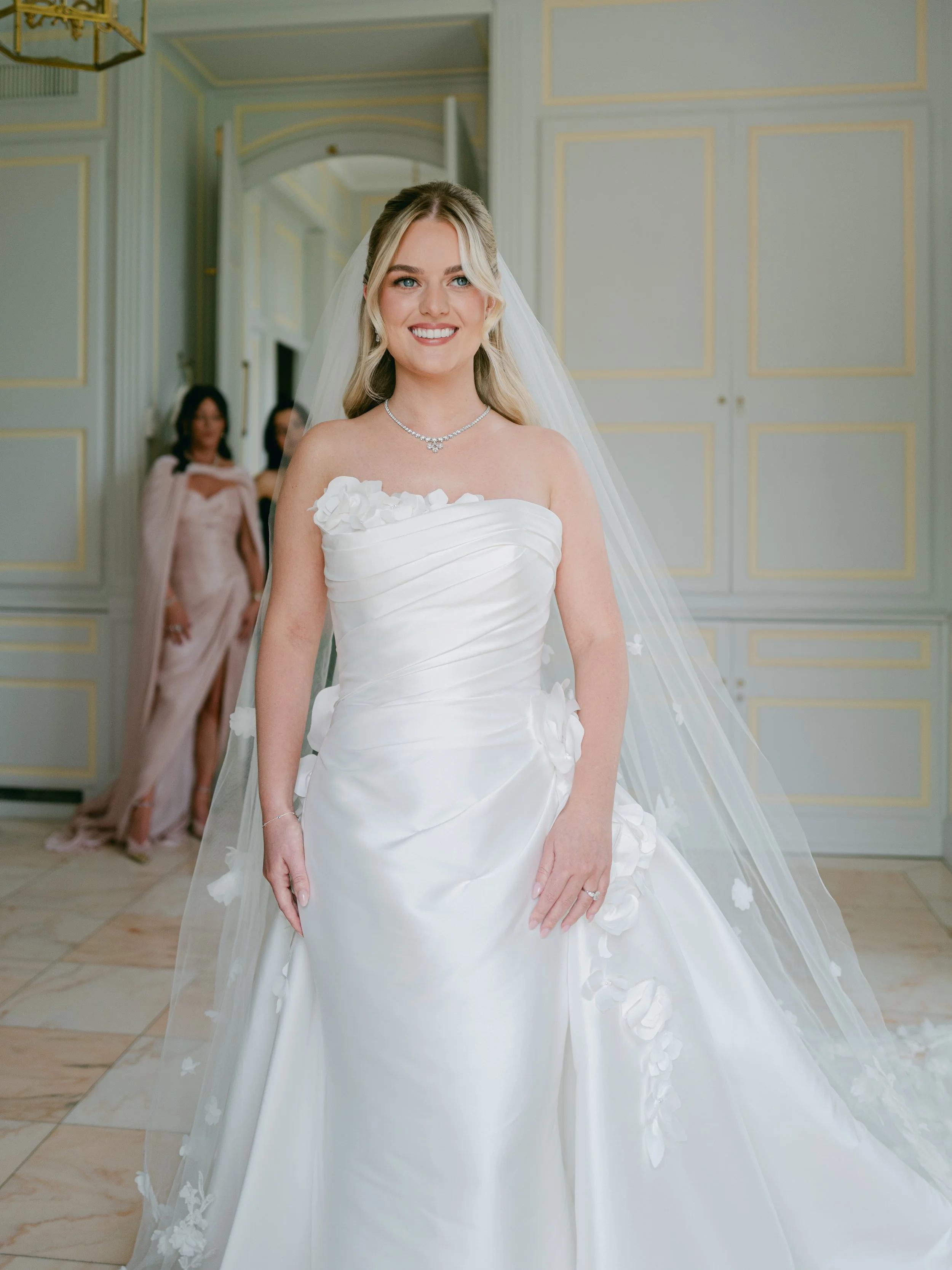 A bride in a white wedding dress with floral accents, wearing a diamond necklace and veil, smiling in a decorated room with gold-trimmed walls and two women in the background.