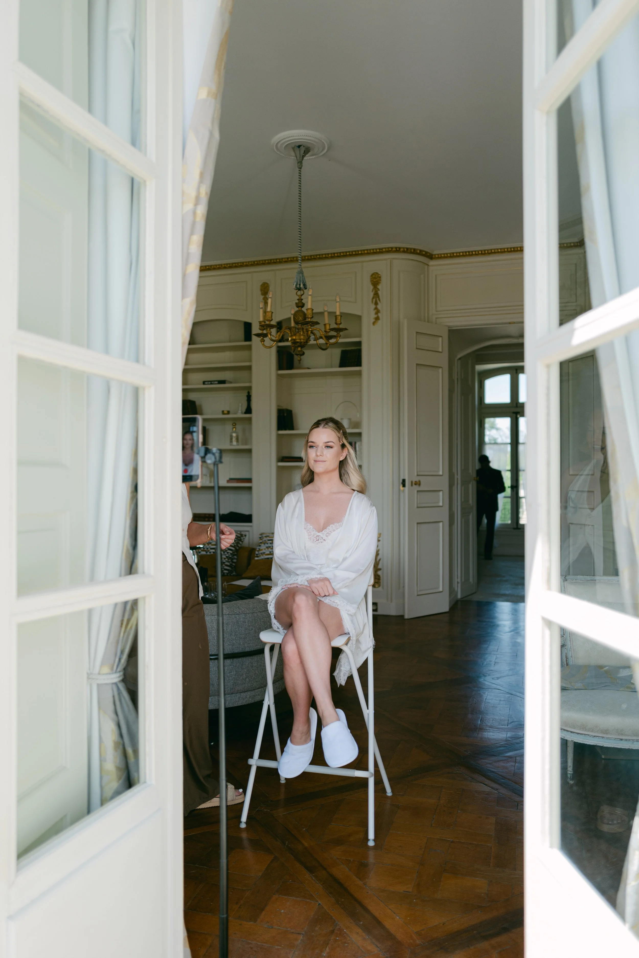 A young woman getting ready, sitting on a chair with crossed legs, wearing a satin robe, in a decorated and elegant room with a chandelier and large windows, as seen through an open French door.