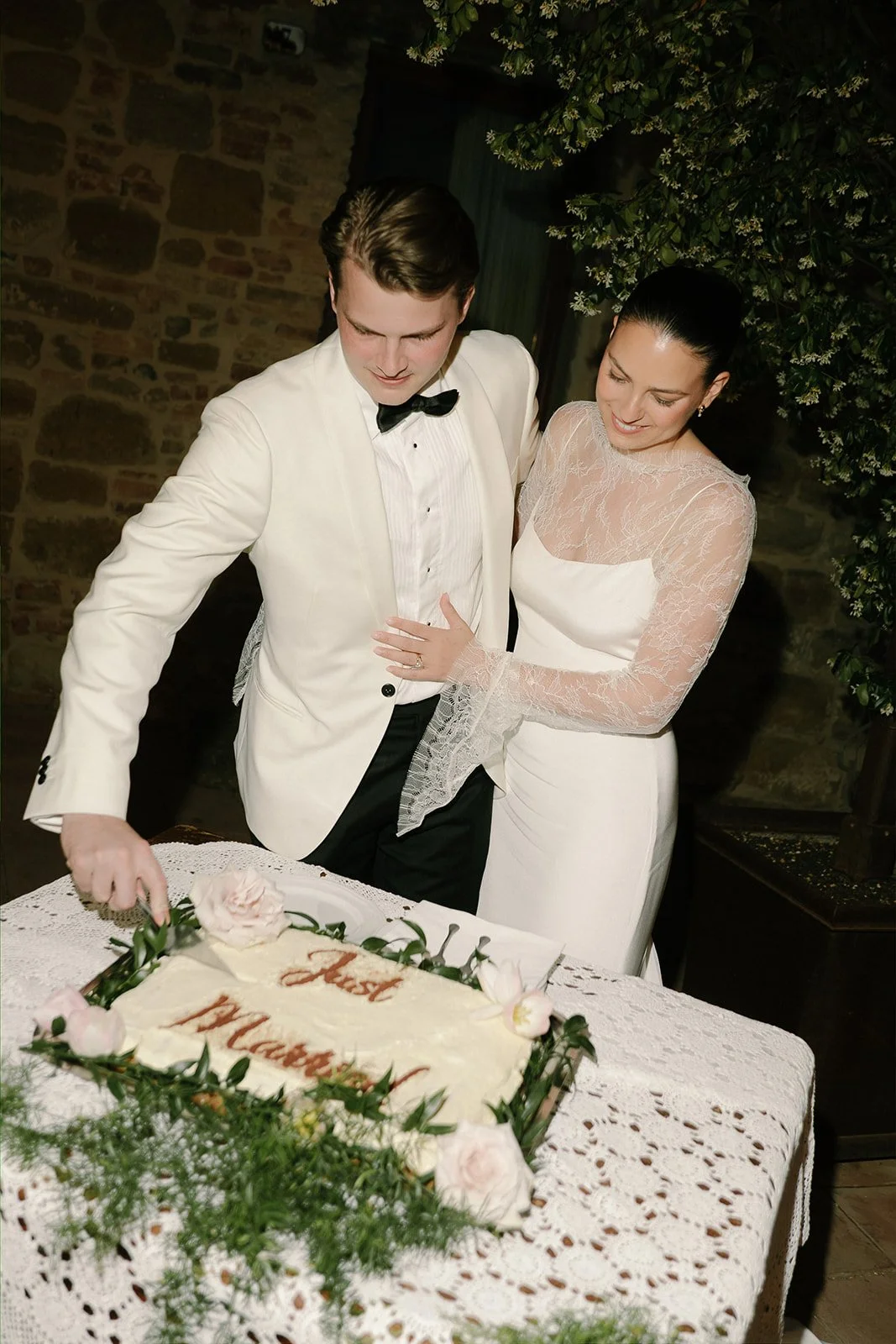 A bride and groom in wedding attire cutting a wedding cake decorated with flowers and greenery, inside a rustic venue with stone walls and greenery.