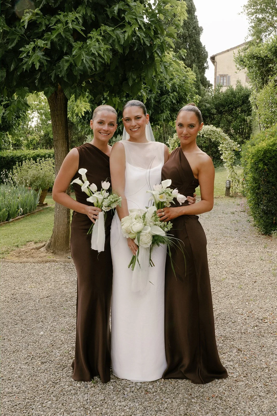 A bride in a white wedding dress standing between two women in brown dresses, holding bouquets of white flowers, outdoors in a garden setting with green trees and plants.