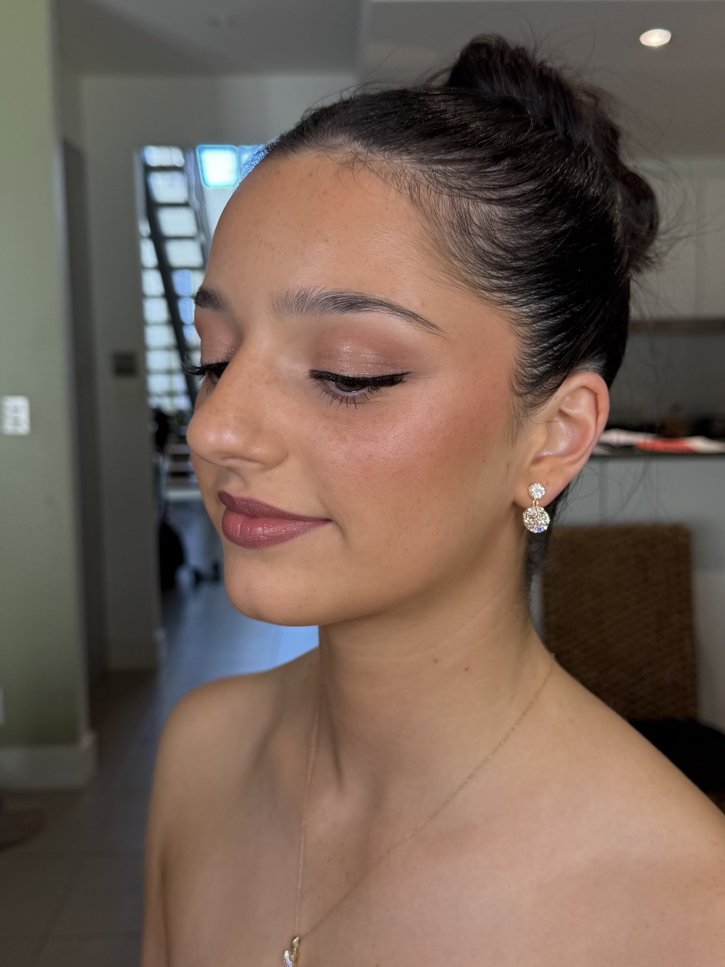 Close-up of a woman's face and upper body, with her eyes closed and wearing earrings and a necklace, indoors with blurred background.