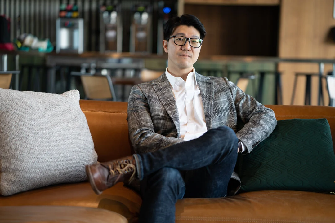 A confident man in glasses, wearing a plaid blazer and white shirt, sits on a brown leather sofa with arms crossed, in a modern cafe or coworking space.