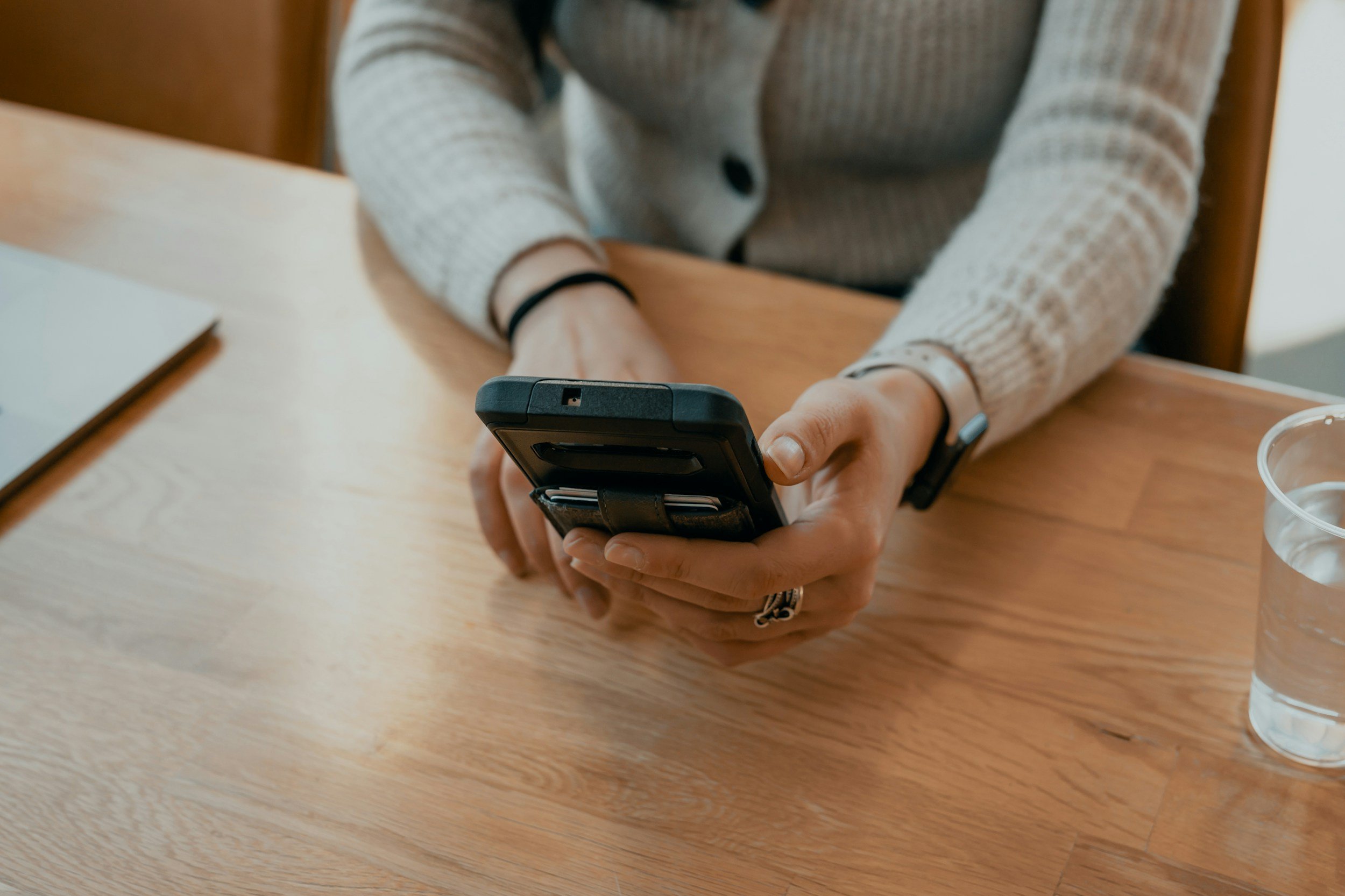 Person sitting at a wooden table, holding a smartphone in both hands, wearing a beige sweater and a black wristband, with a glass of water on the side.