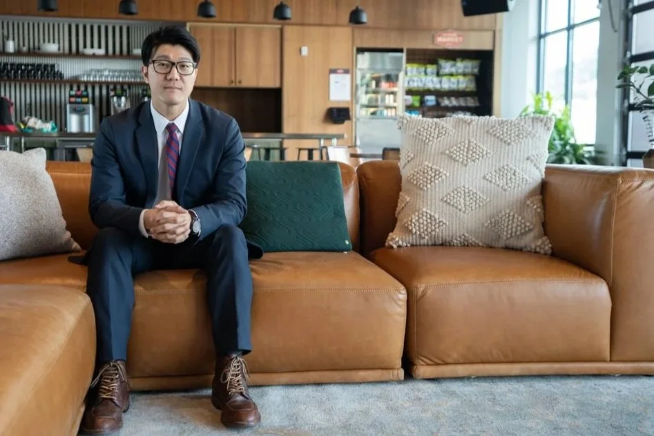 A man in a navy suit, white shirt, and striped tie sitting on a tan leather couch in a modern cafe or lounge with large windows, wooden shelving, and plants in the background.