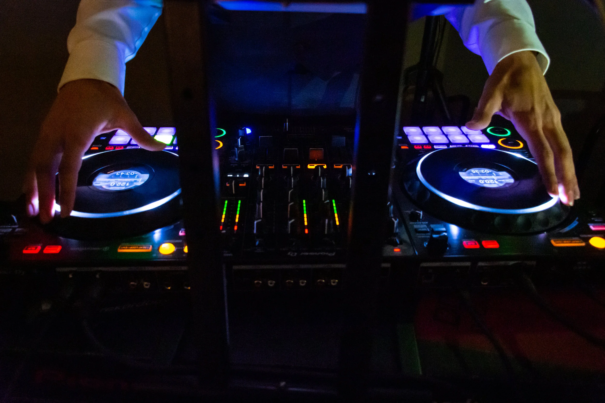 A DJ performing with two turntables and a mixer in a dark environment, with colorful LED lights reflected on the equipment.