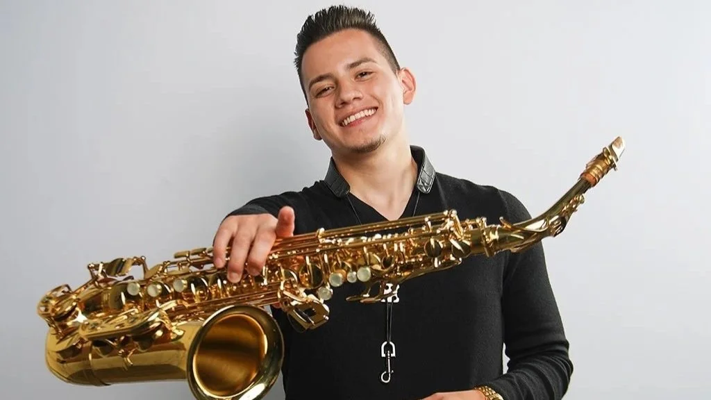 A young man with short dark hair and a light beard, smiling and wearing a black shirt, holding a gold-colored saxophone in front of a plain white background.
