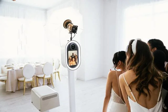 People at a photo booth with a camera and screen, in a bright white room decorated for a celebration or event.