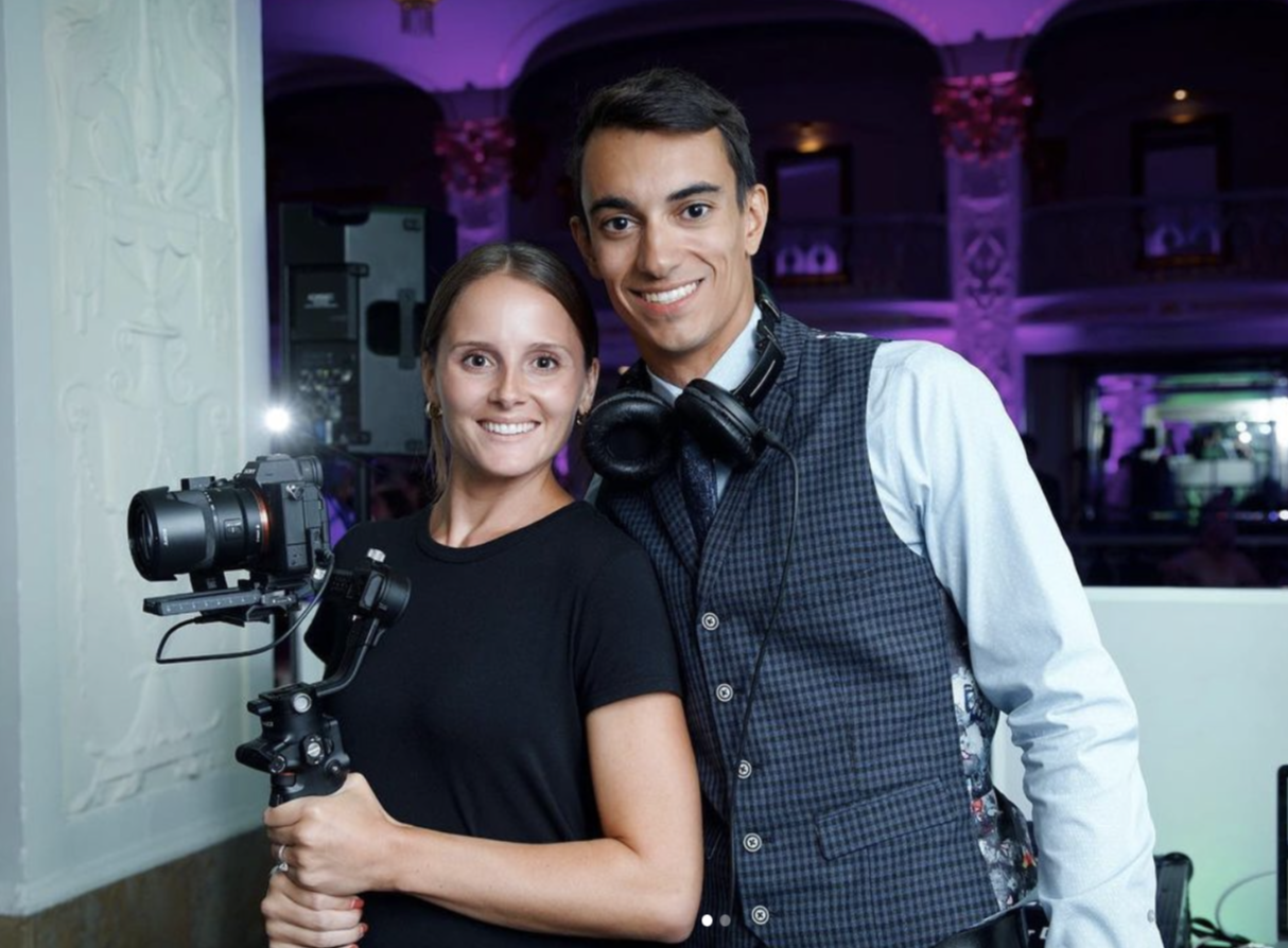Two young adults, a woman with a camera and a man with headphones, smiling at a party or event with purple lighting and ornate decor in the background.