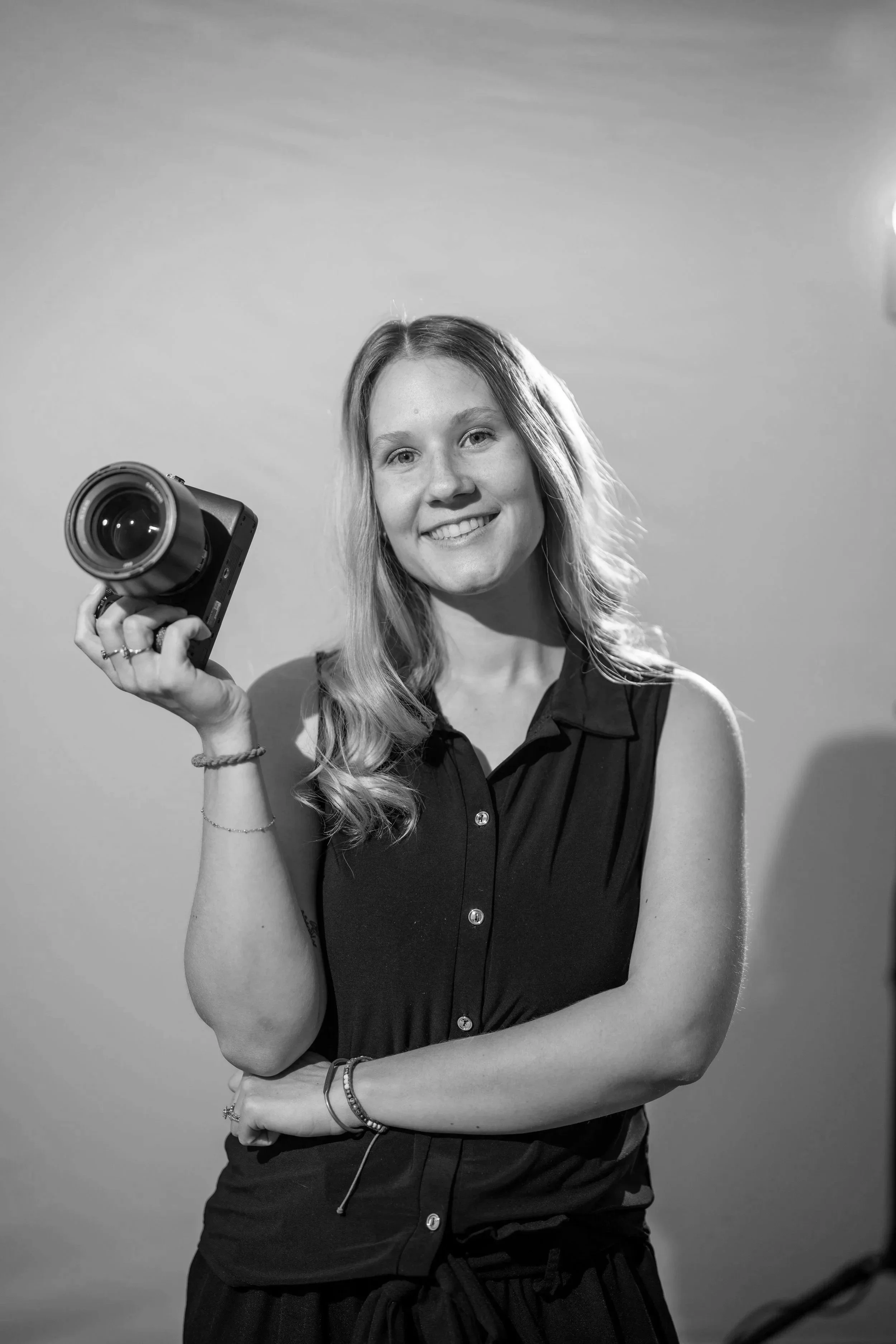 A young woman with long hair smiling and holding a camera, standing against a plain background in black and white.