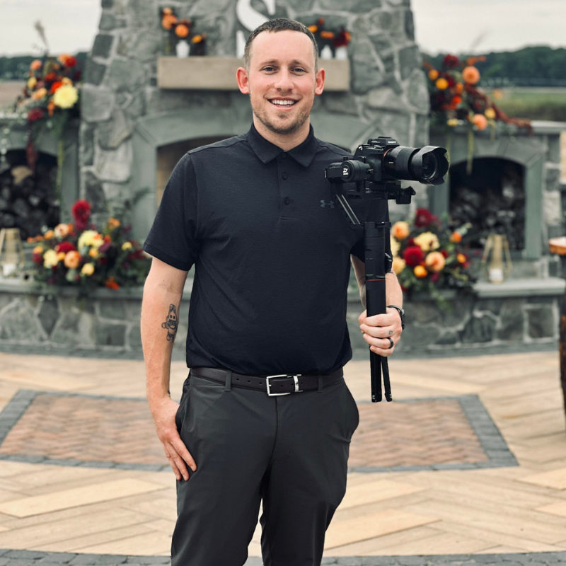 A man in a black polo shirt and dark pants holding a camera on a tripod in front of a stone fireplace decorated with orange, yellow, and red flowers outdoors.
