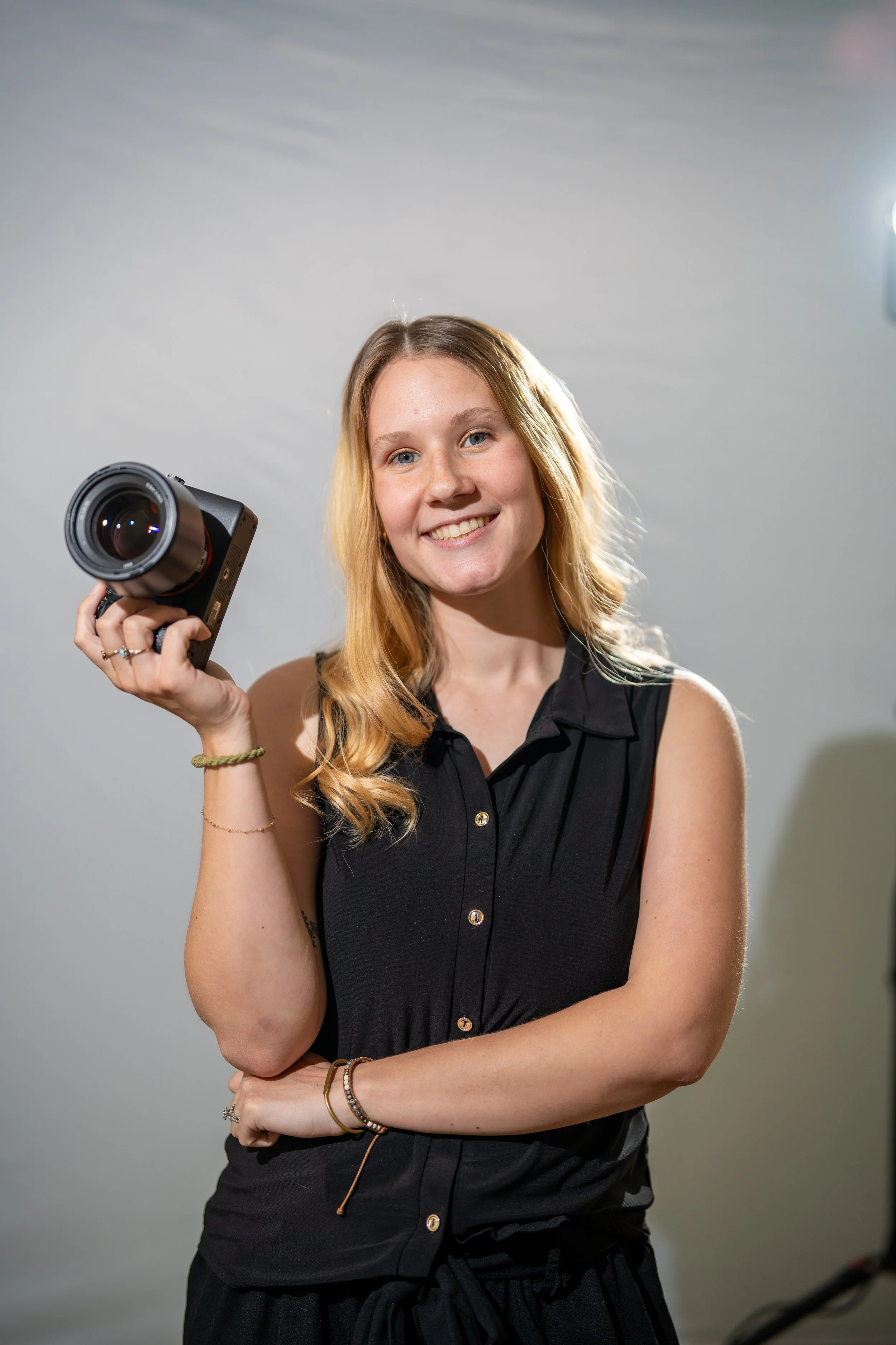 Young woman with long, wavy blonde hair smiling playfully, holding a professional camera with a large lens, wearing a black sleeveless button-up top, standing against a plain gray background.