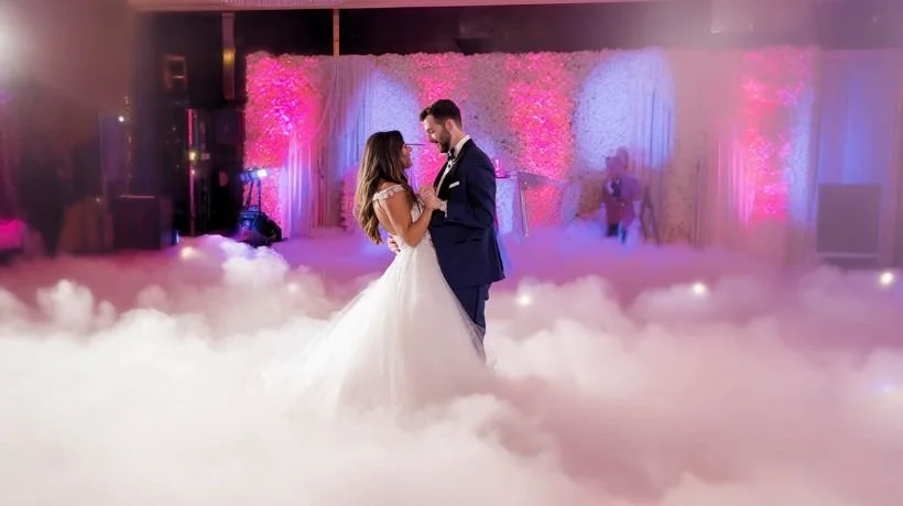 A bride and groom dancing on a fog-filled dance floor at their wedding reception with pink and purple lighting and floral decorations in the background.
