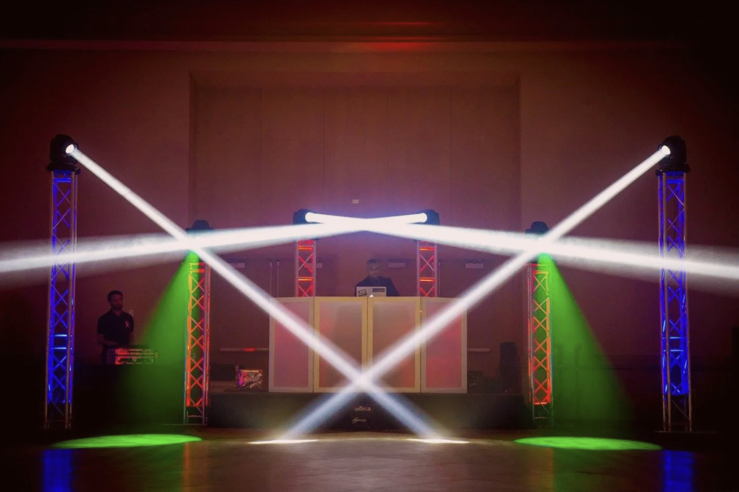 Stage with colorful lighting, laser beams crossing, DJ booth in the center, and two people managing equipment.