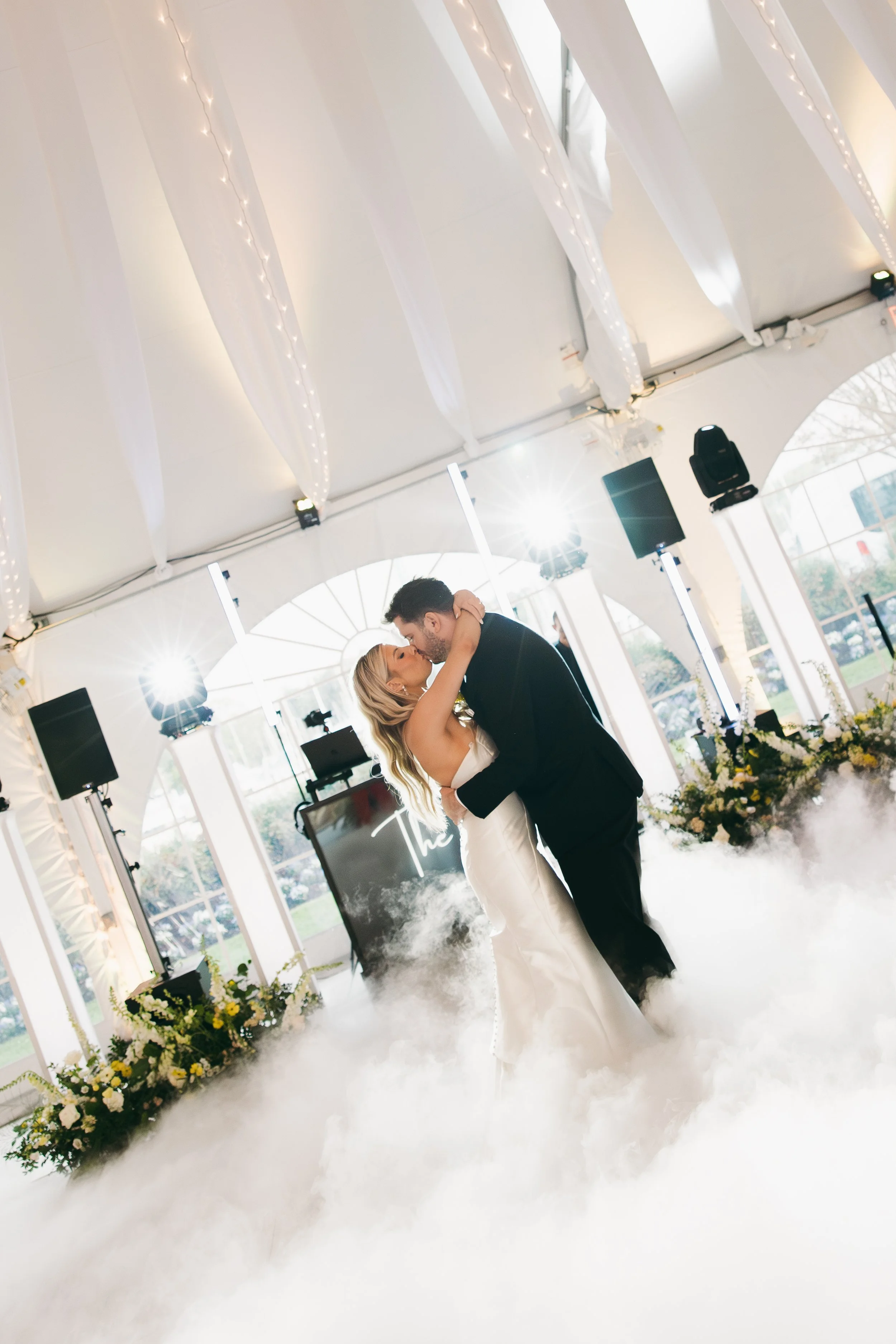 A bride and groom share a first dance in a decorated wedding reception tent with white drapery, string lights, and floral arrangements, with fog on the floor.