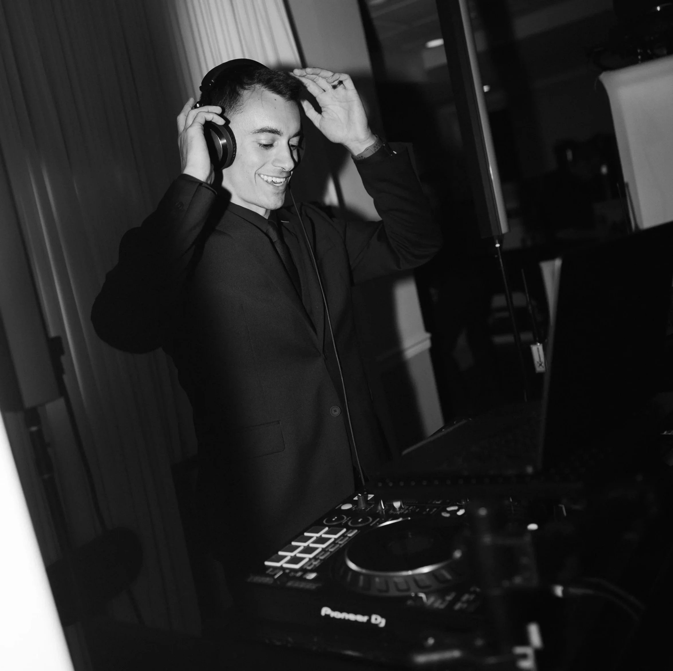 DJ Topedo dressed in a suit is smiling while wearing headphones and standing behind a DJ mixer, adjusting his headphones with one hand.