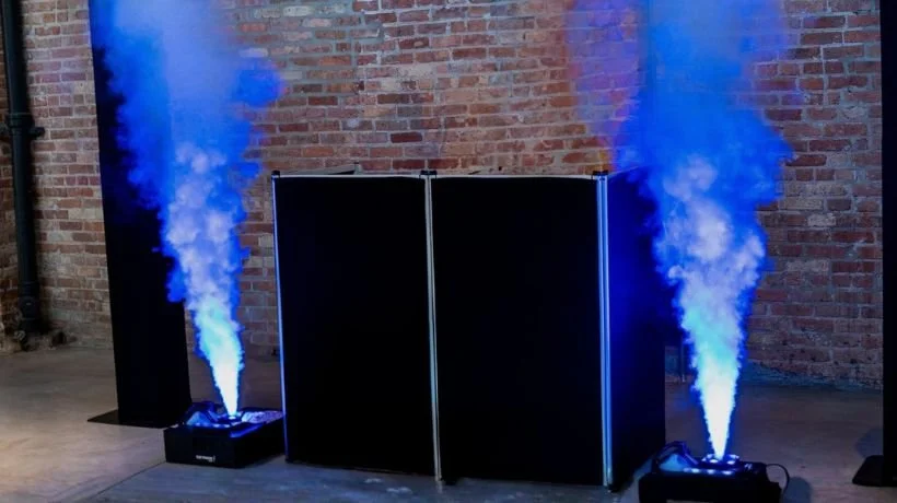Stage setup with blue smoke machines emitting blue smoke in front of black folding screens against a brick wall.