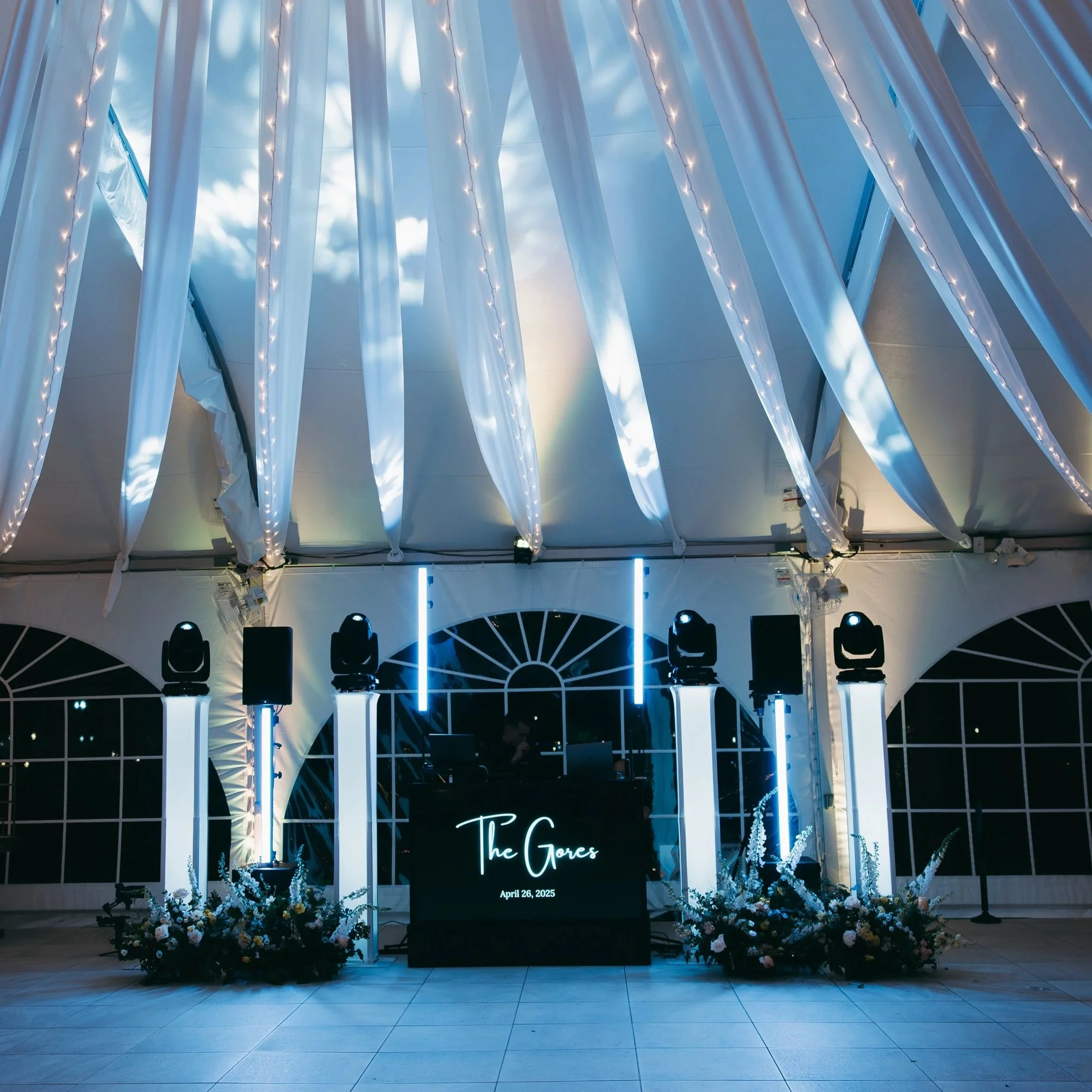 Elegant event setup inside a tent featuring a DJ booth with a sign reading 'The Gores' and the date April 26, 2025. The ceiling is decorated with white drapes and string lights, with colorful lighting and floral arrangements at the base of the columns.