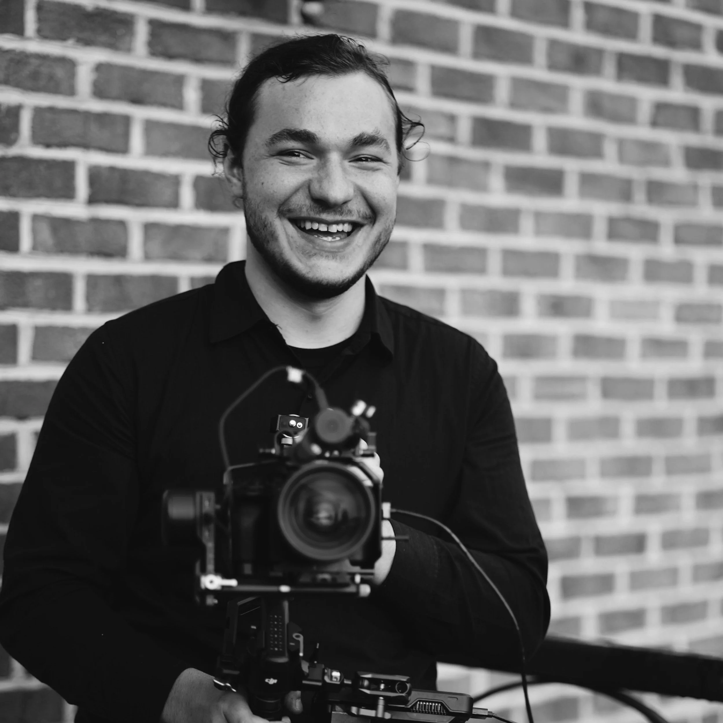 A smiling man holding a professional camera in front of a brick wall.