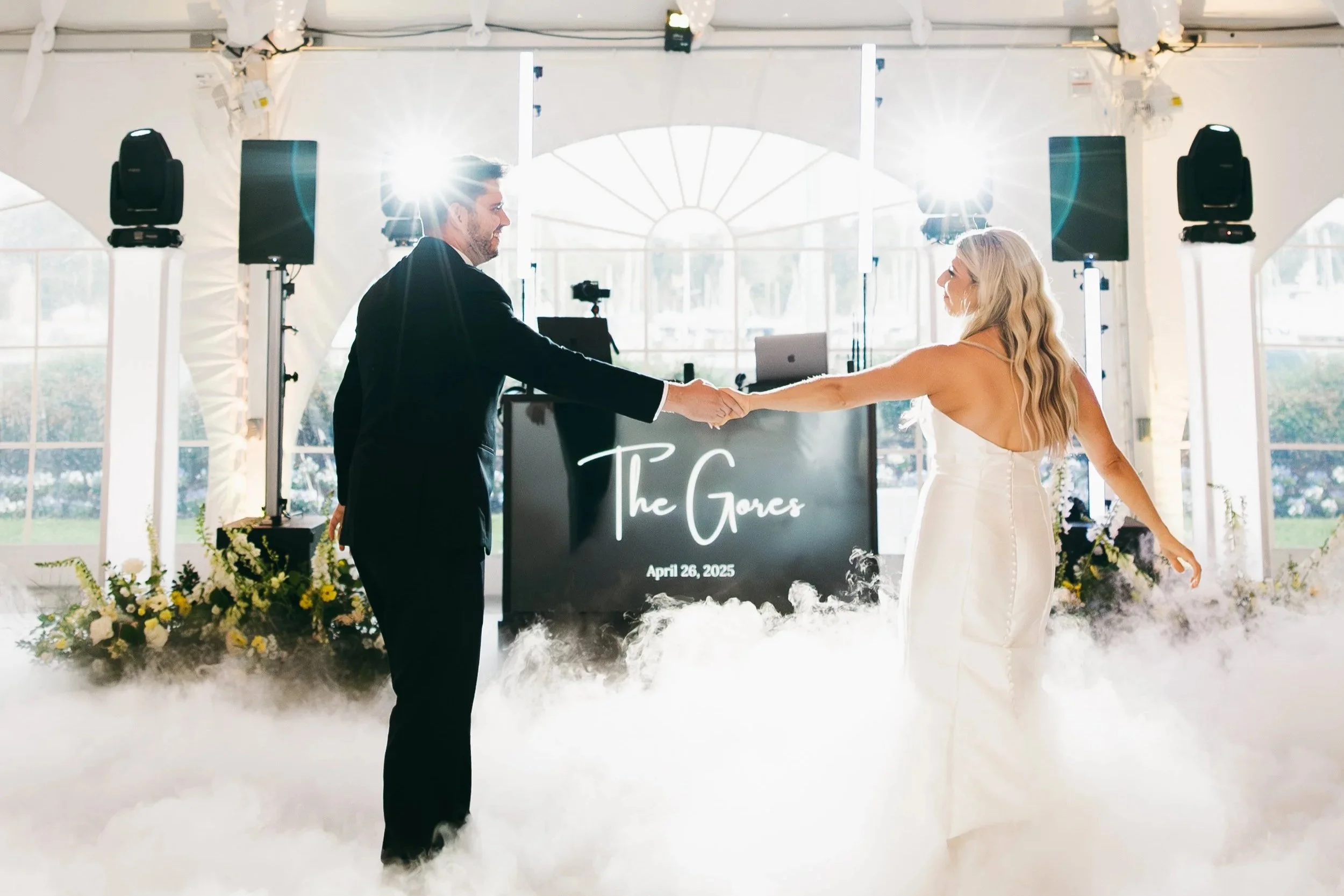 A newlywed couple, a man in a black suit and a woman in a white wedding dress, holding hands and dancing at their wedding reception in a bright, decorated venue with fog on the floor. A sign with the names 'The Gores' and the date 'April 26, 2025' is visible in the background.
