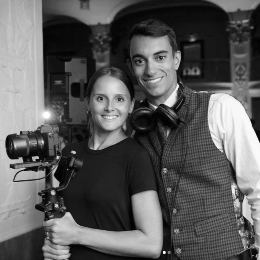 A woman and a man smiling for a photo, with the woman holding a camera with a stabilizer. They are indoors, in a room with decorative columns, and both are dressed casually.