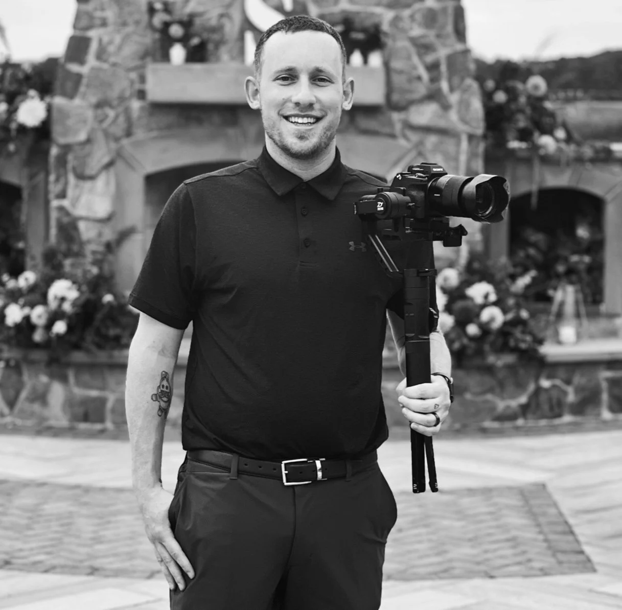 A man smiling and holding a camera on a tripod outdoors, in front of a stone fireplace decorated with flowers