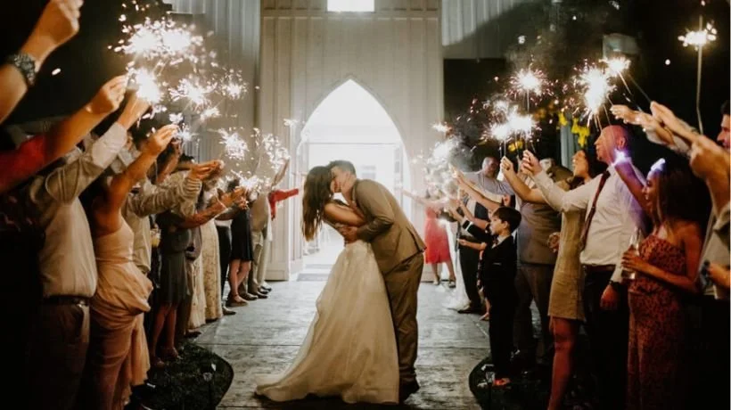 A bride and groom share a kiss under a canopy of sparklers held by wedding guests at their wedding reception.