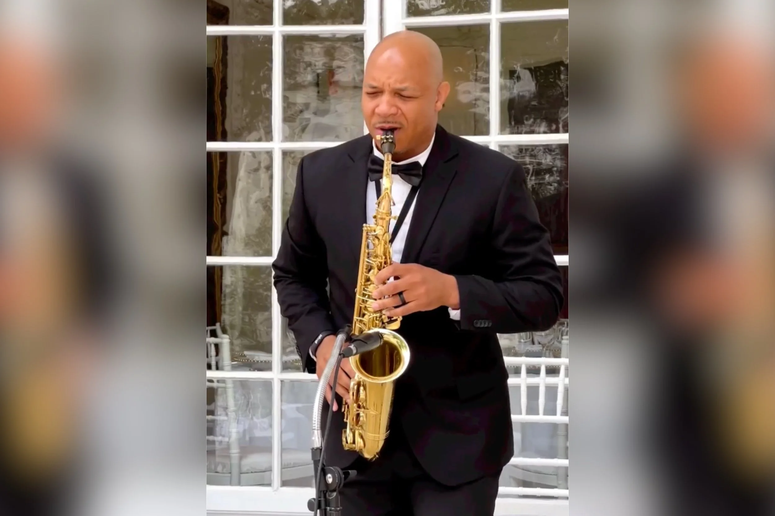 A man in a black tuxedo and bow tie playing a gold-colored saxophone outdoors in front of a windowed building.