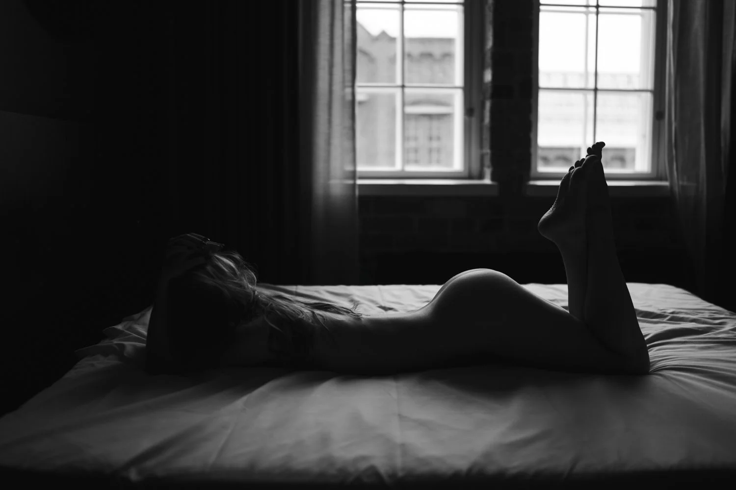 Mustavalkoinen taiteellinen alastonkuva, vastavalo korostaa vatsallaan makaavan naisen vartalon kaarta.