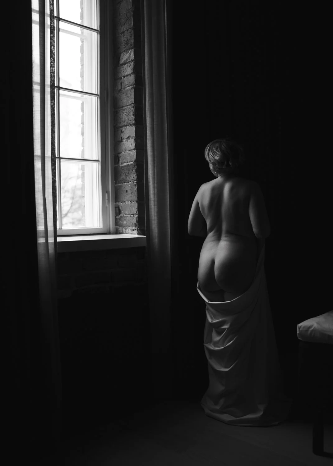 Nainen poseeraa ikkunan edessä pelkkään lakanaan kietoutuneena boudoir-kuvauksessa.