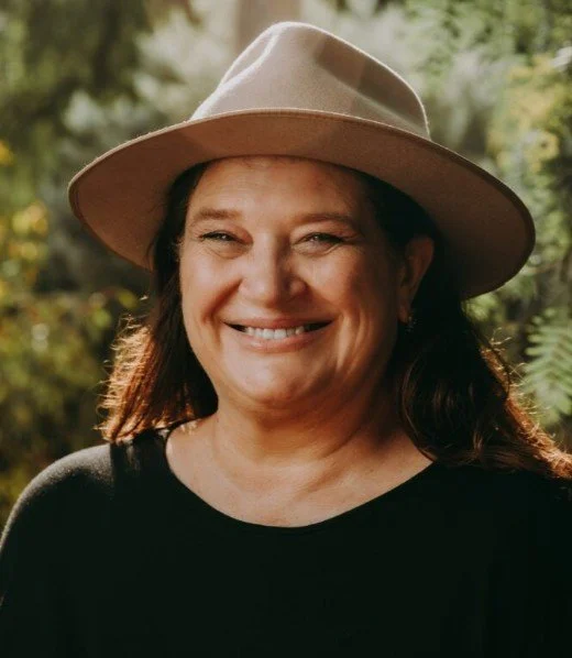 Woman smiling outdoors, wearing a beige wide-brim hat and a black top, with blurred greenery in the background.
