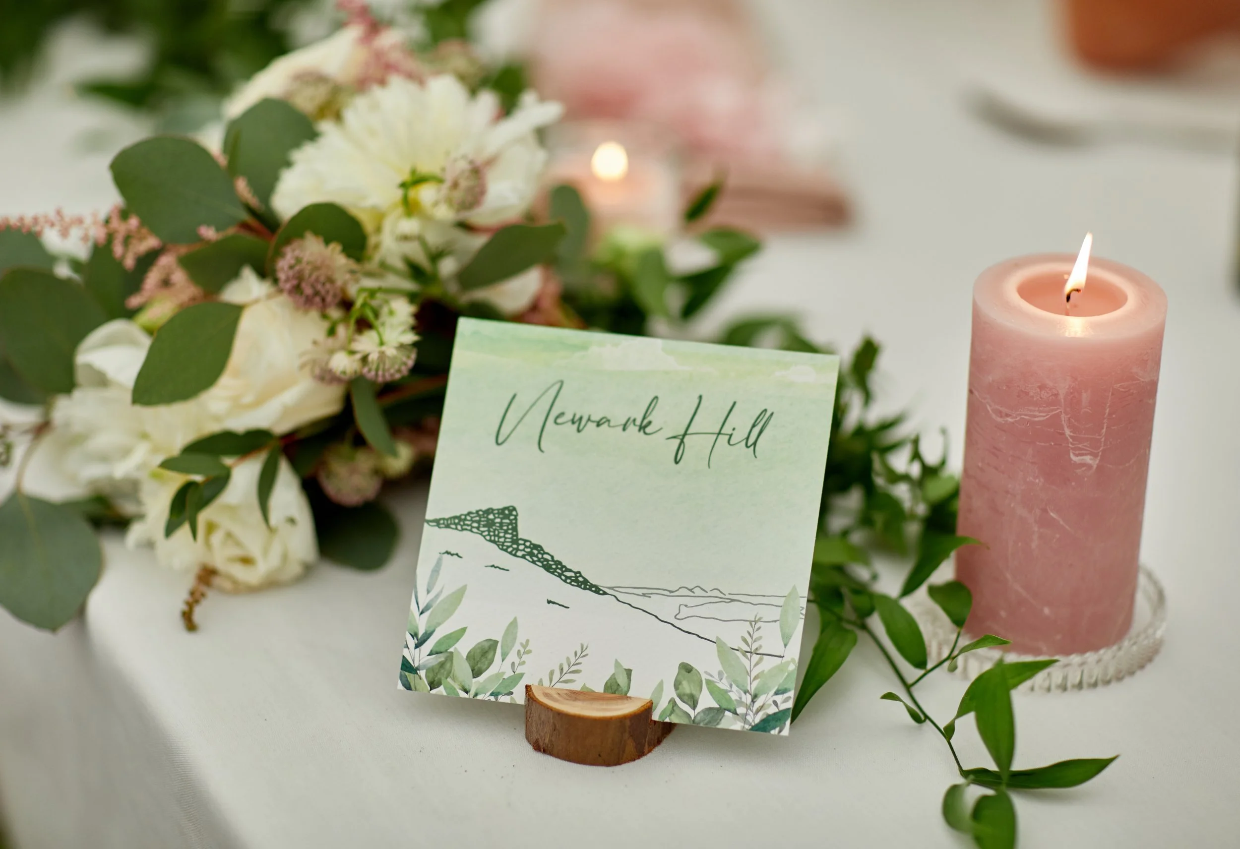 A wedding table name card featuring a hand-drawn illustration of a landmark from a map set on a table with white and green foliage and a pink candle