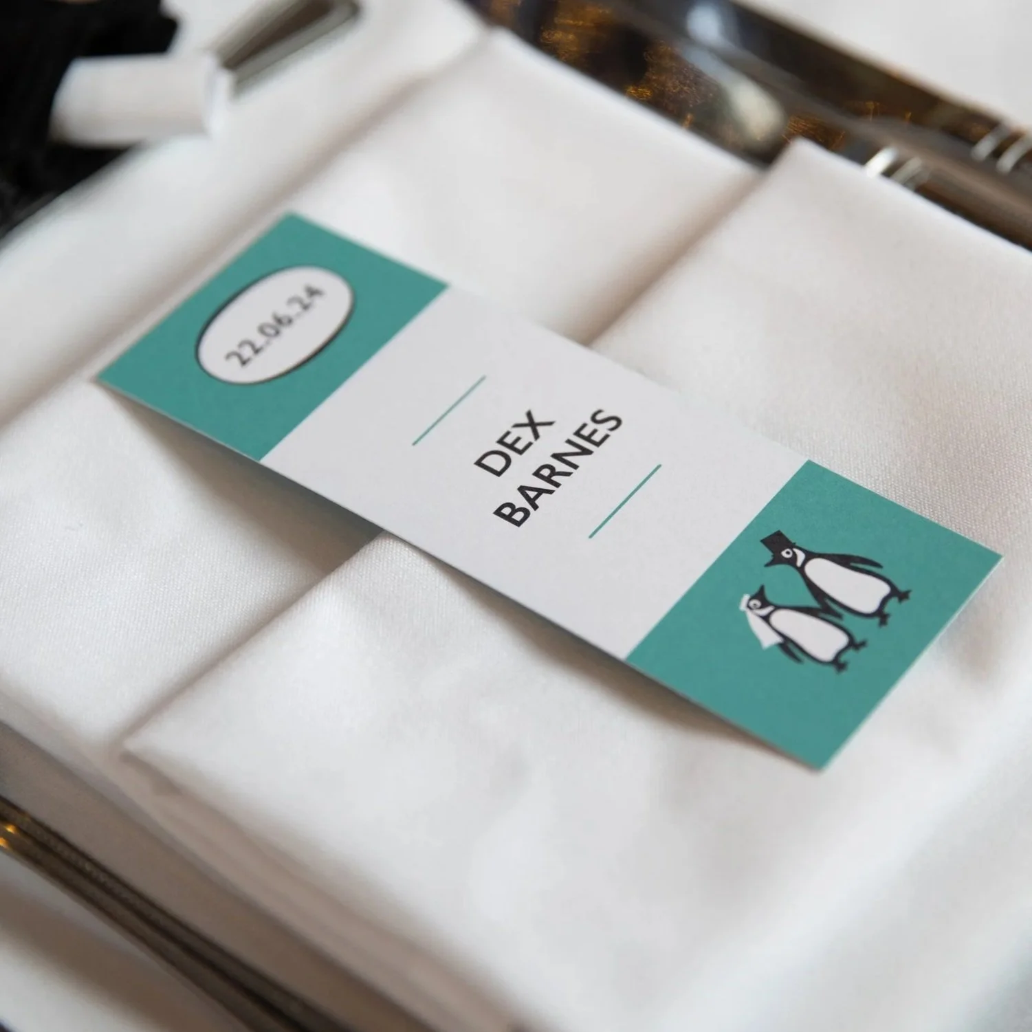 An teal and white bookmark wedding place card styled as a classic penguin book sits on a napkin on a place setting at a wedding table.