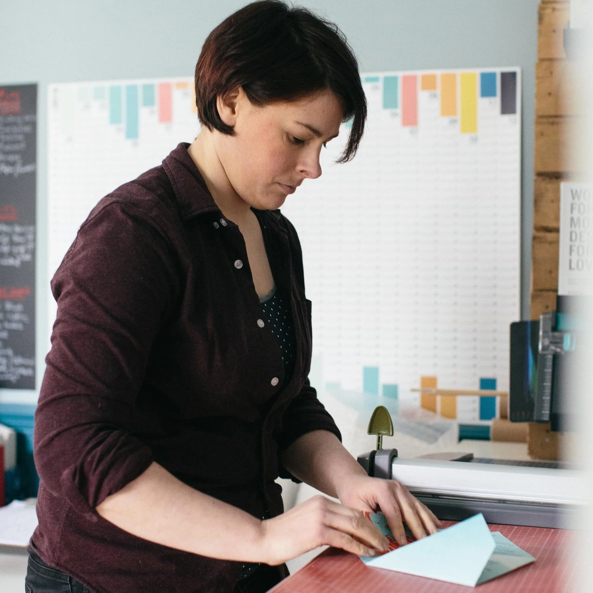 A white, scottish female with dark brown hair, wearing a burgundy shirt, starts at a cutting mat folding an origami wedding invitation. There are colourful wall planners on the walls of the room behind her.
