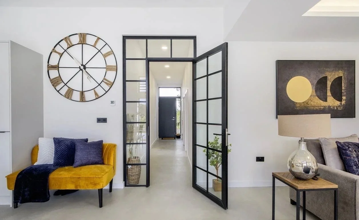 Modern living room interior with a large wall clock, yellow sofa with cushions, glass-panel door, abstract wall art, and side table with lamp.
