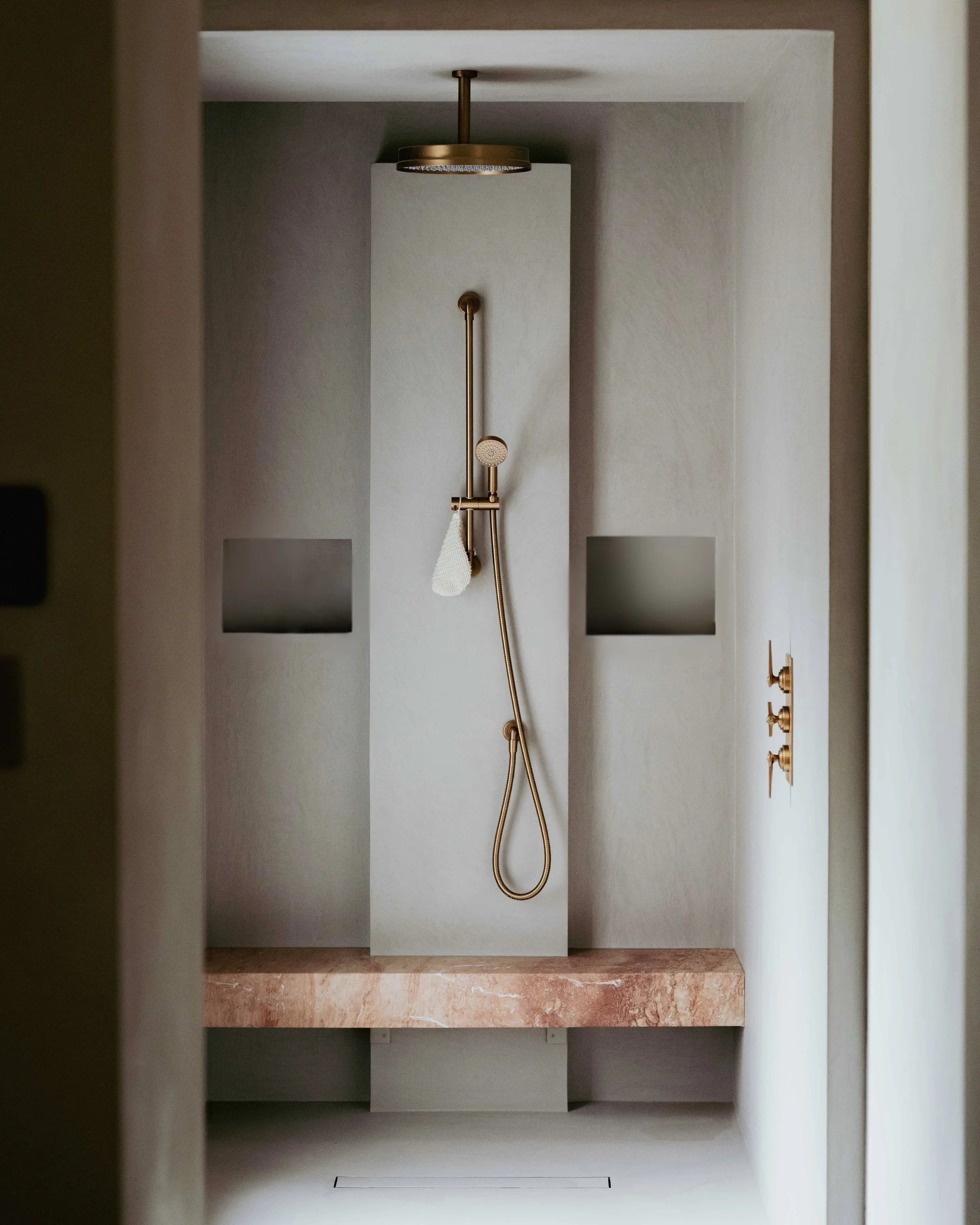Minimalist walk-in shower with gold fixtures, a wall-mounted shower head, and a pink marble bench.
