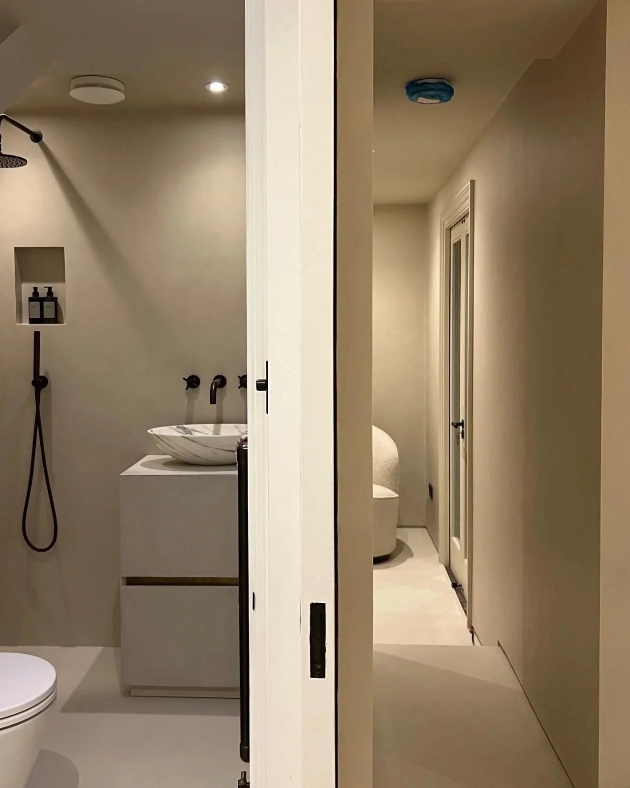 Microcement bathroom with seamless beige walls, modern black fixtures, and a minimalist hallway view