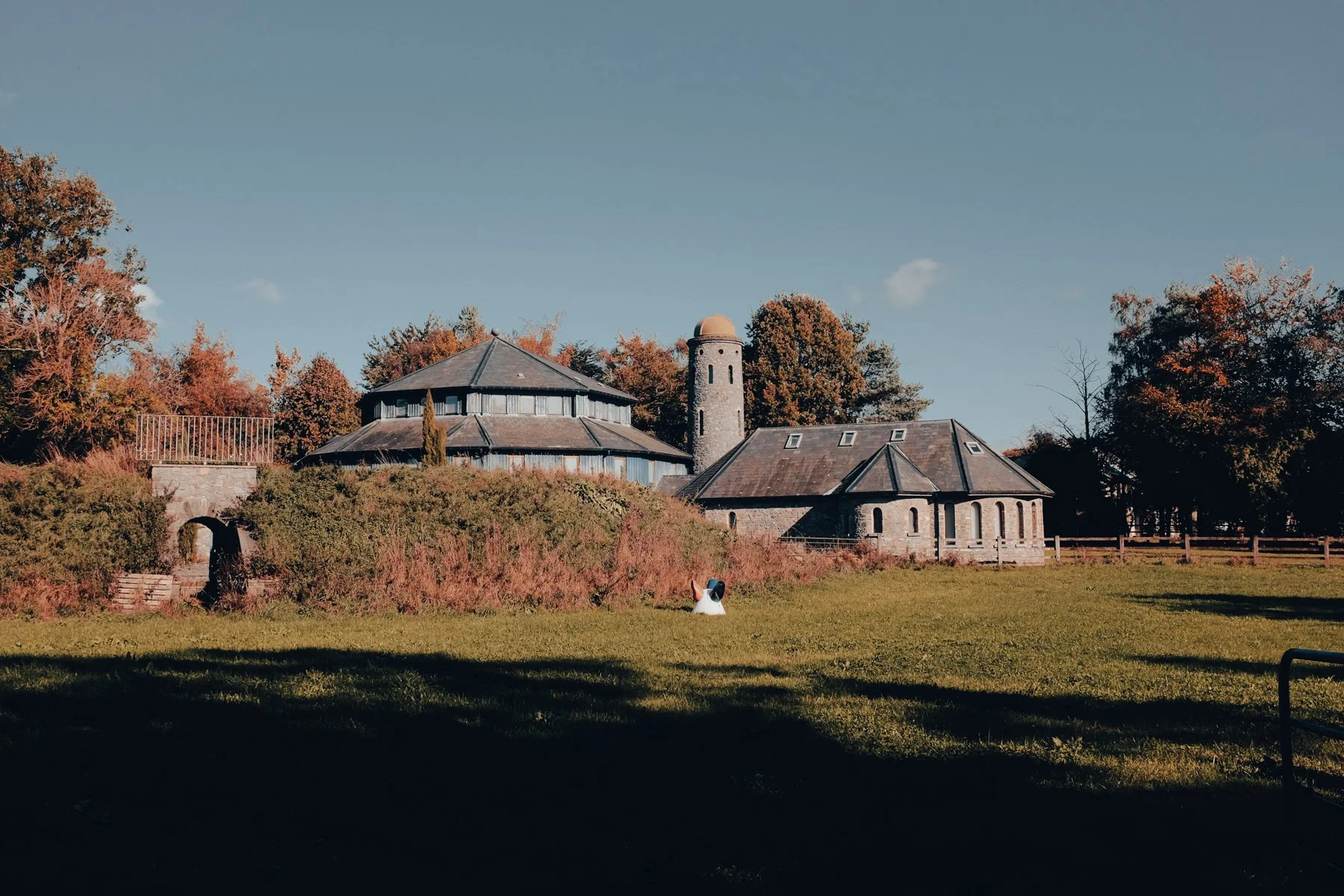 Stone building with round tower and sloped roofs, surrounded by grassy field and trees, under a clear blue sky.