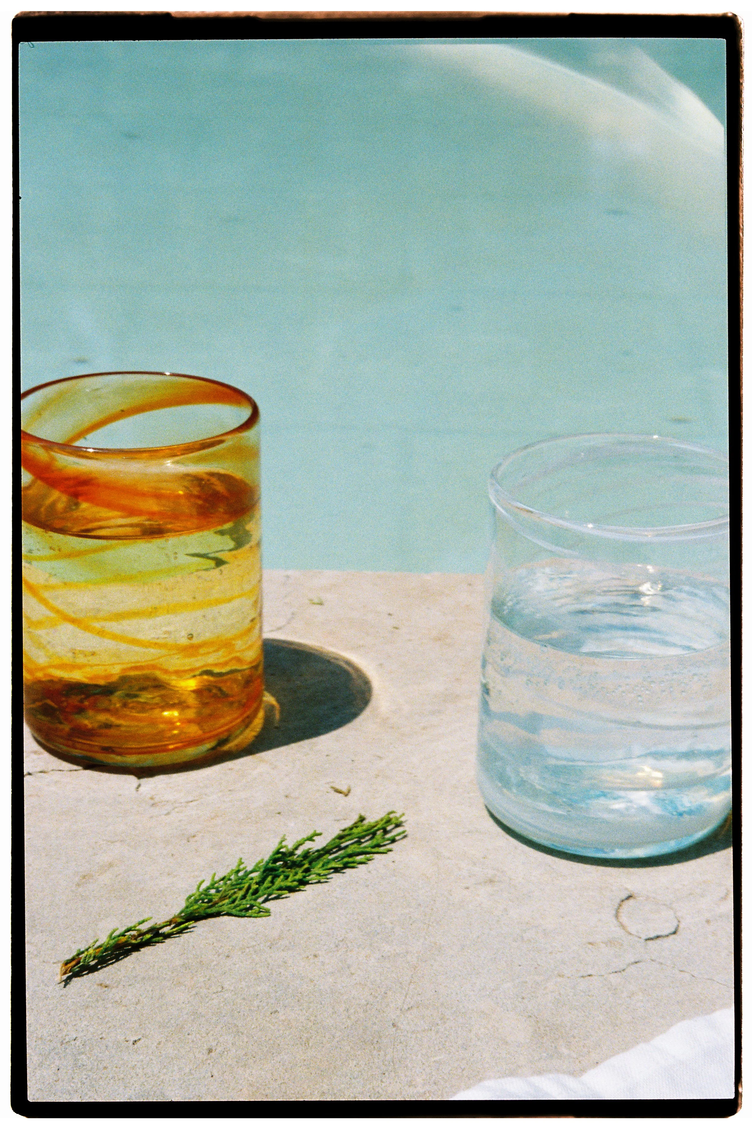 Two glasses of water, one with an amber liquid, on a poolside surface with a sprig of rosemary in front, and a clear swimming pool in the background.