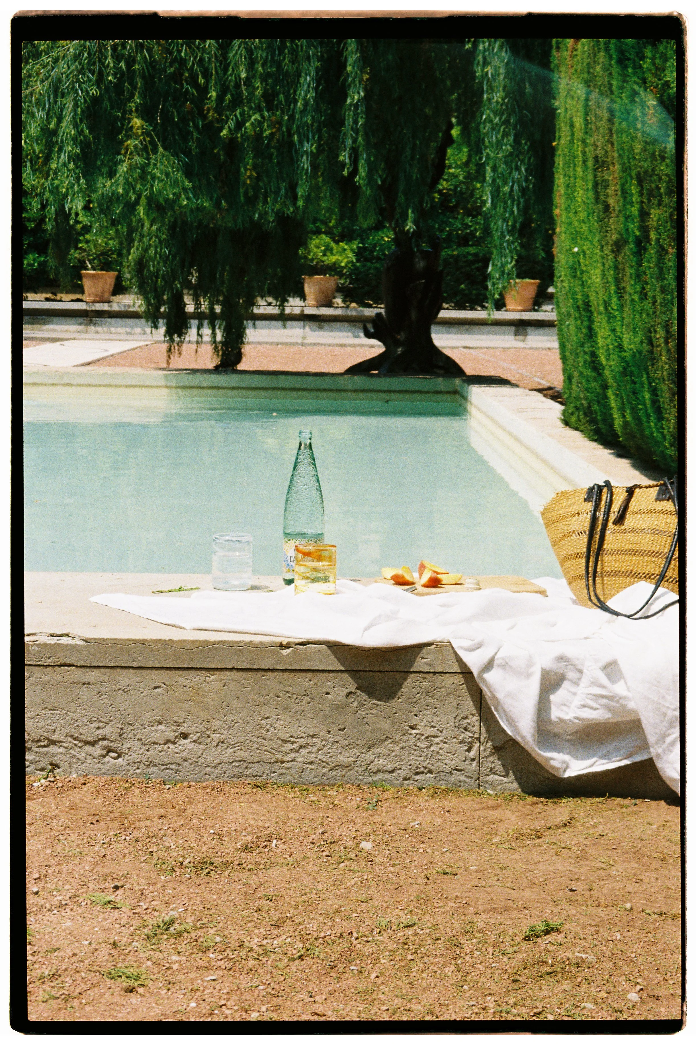 A table with a white cloth on a poolside, holding a glass bottle, glass, apples, and a woven straw bag, near a swimming pool with lush green trees in the background.