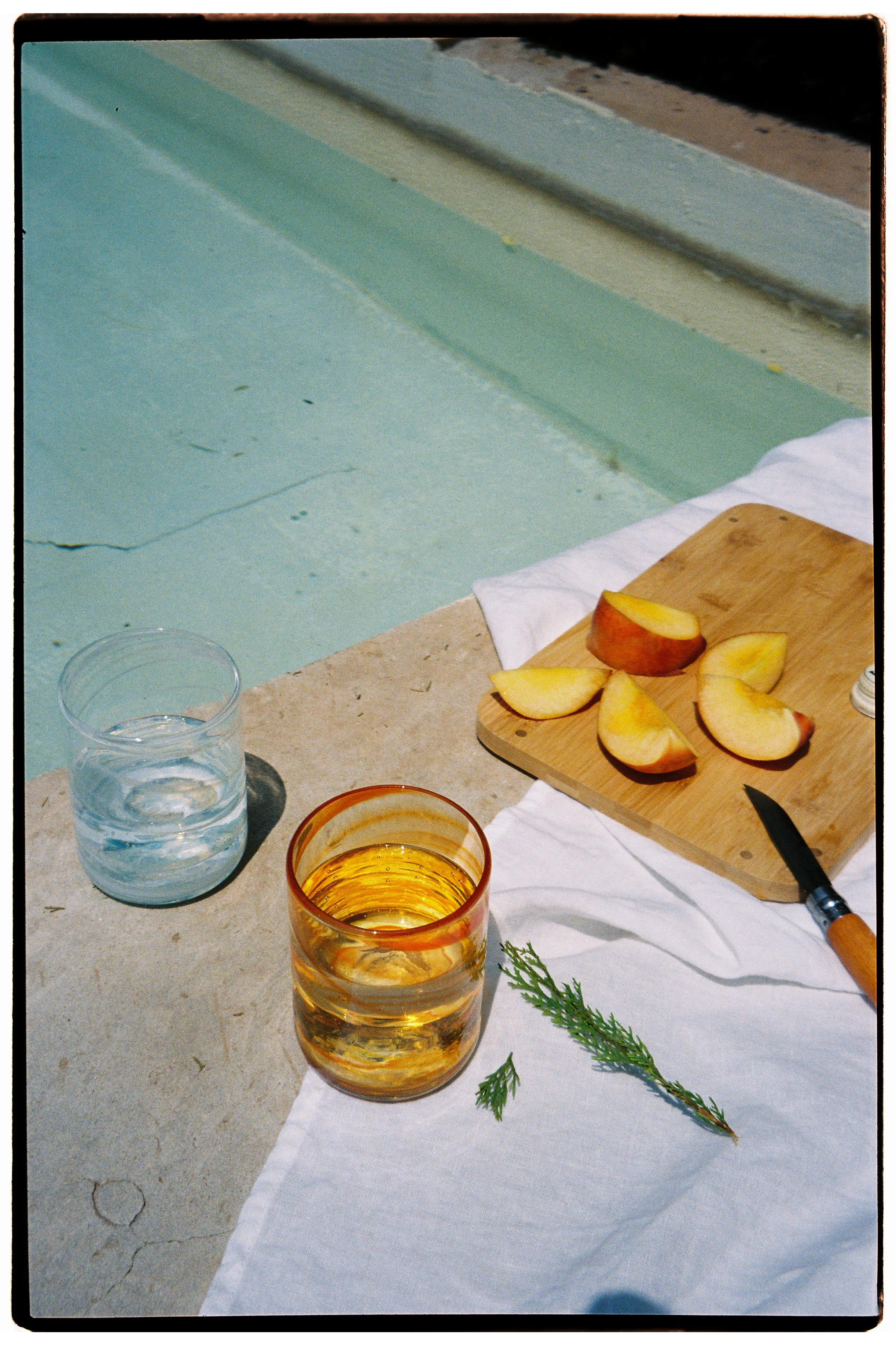 A scene by a swimming pool featuring two glasses of water and a drink with ice, a wooden cutting board with sliced peaches, a black-handled knife, and green herbs, all arranged on a white cloth on a concrete surface.