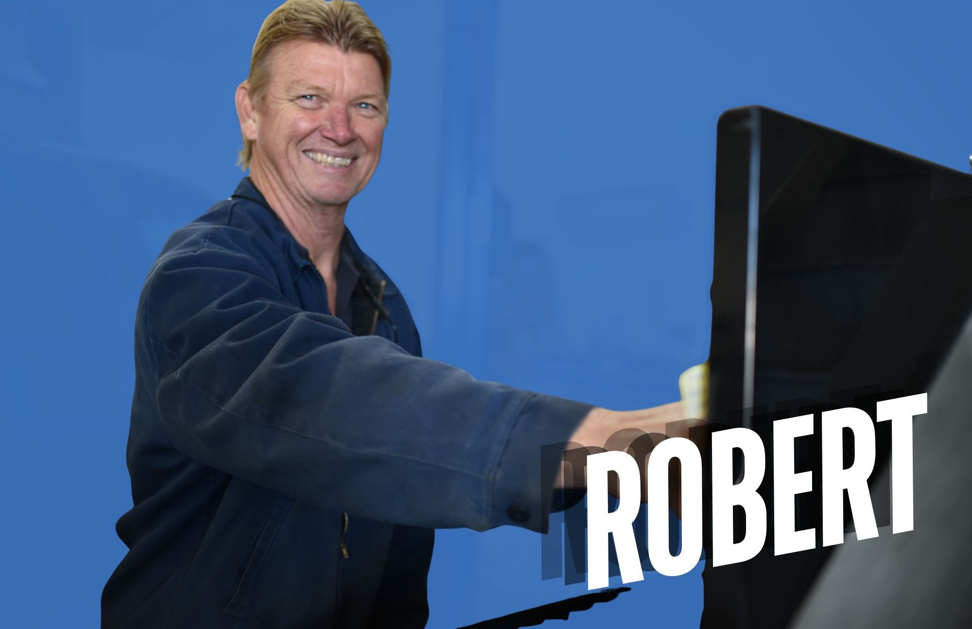A smiling man with blonde hair in a blue shirt, reaching out to touch a sign that reads 'ROBERT' against a blue background.