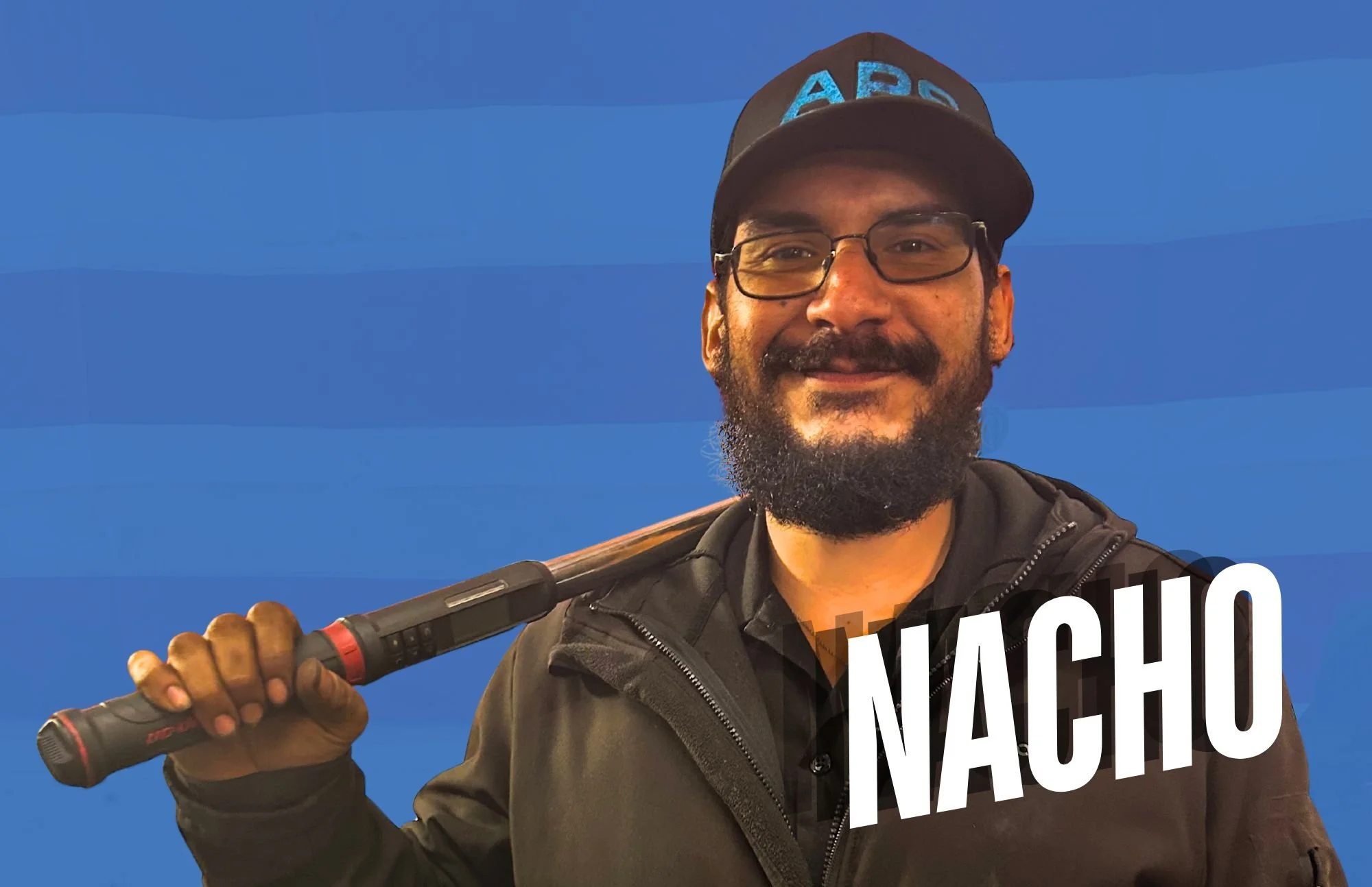 A man with glasses, a beard, and a cap with blue letters, smiling and holding a baseball bat over his shoulder against a blue background, with the name 'Nacho' written in large white letters.