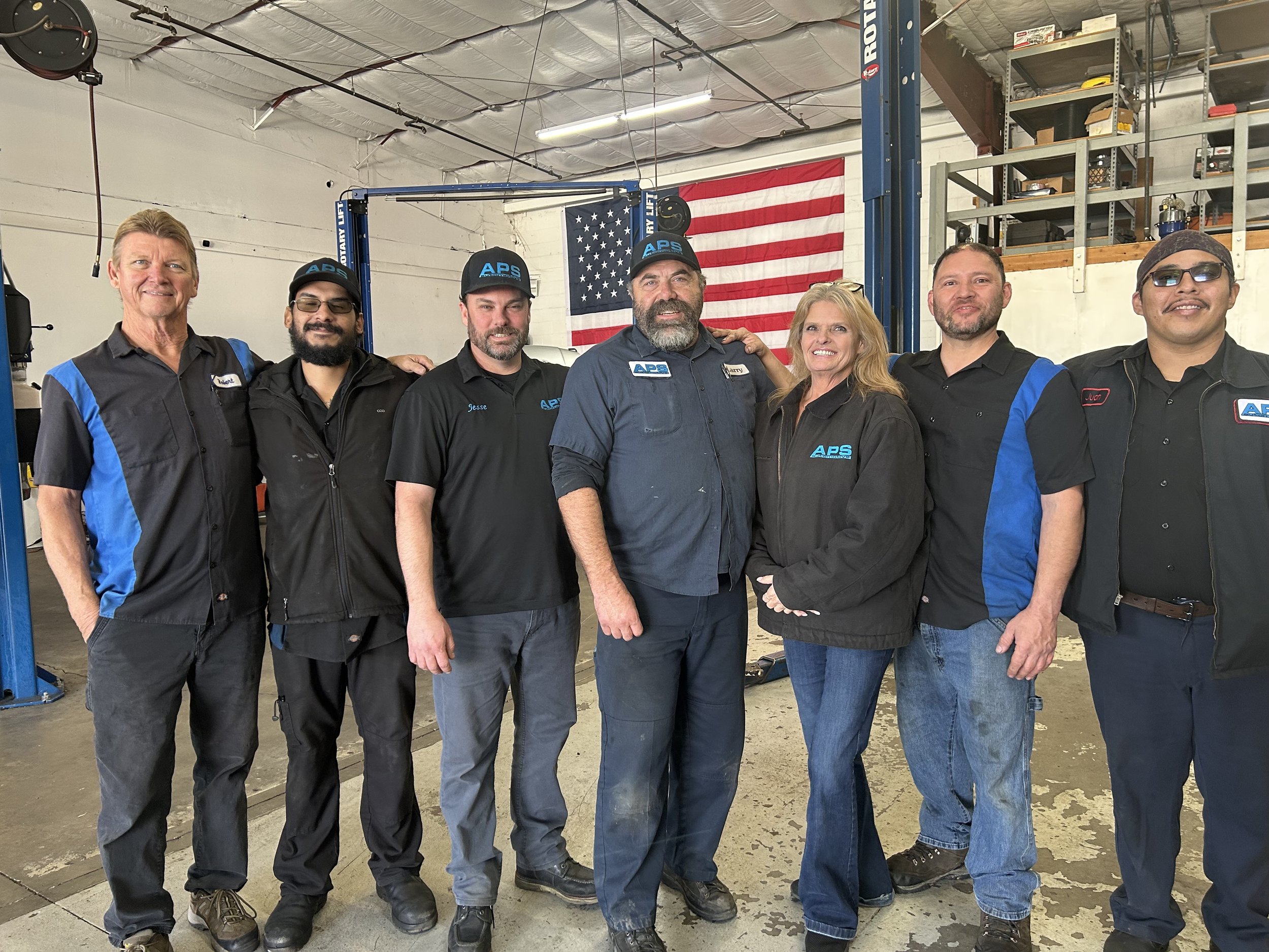 Group of seven people standing inside an industrial workshop, smiling at the camera, with American flag hanging on the wall behind them.