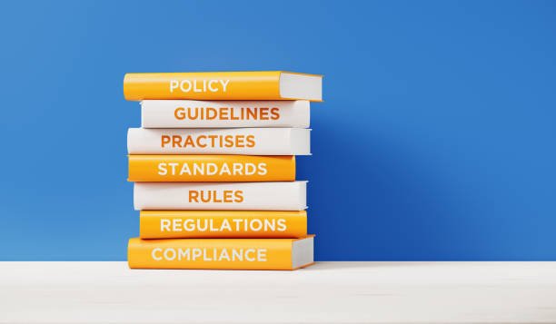 Stack of seven books with titles policy, guidelines, practises, standards, rules, regulations, compliance, on a white surface against a blue background.