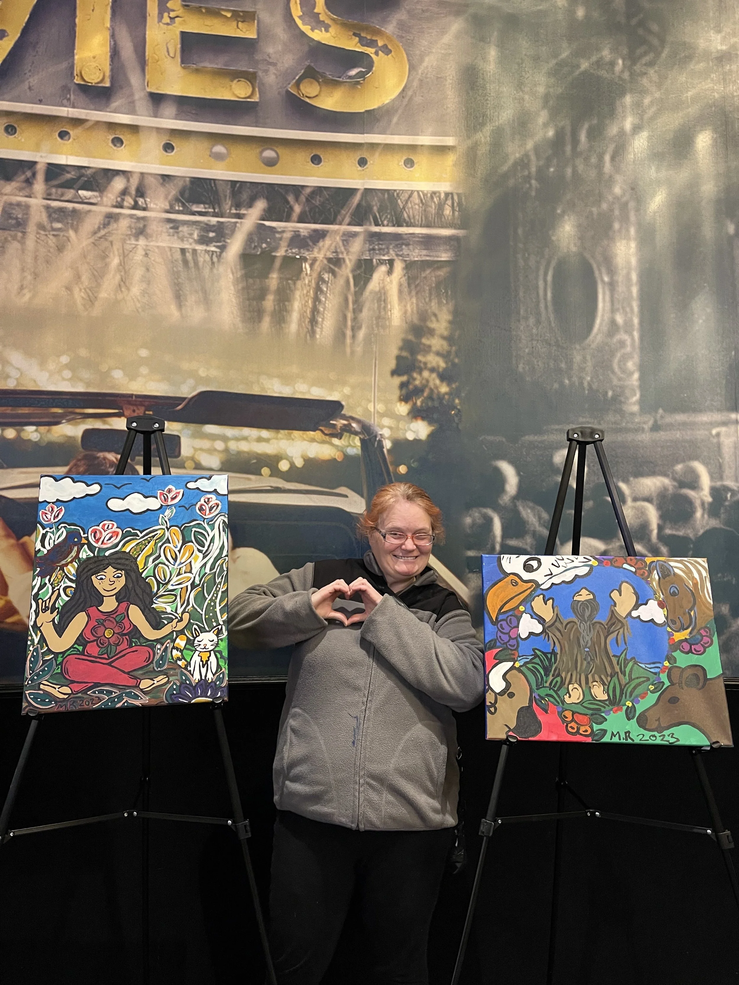 A Deaf autistic woman smiles and forms a heart shape with her hands while standing between two colorful paintings displayed on easels, representing healing and self-expression through art.