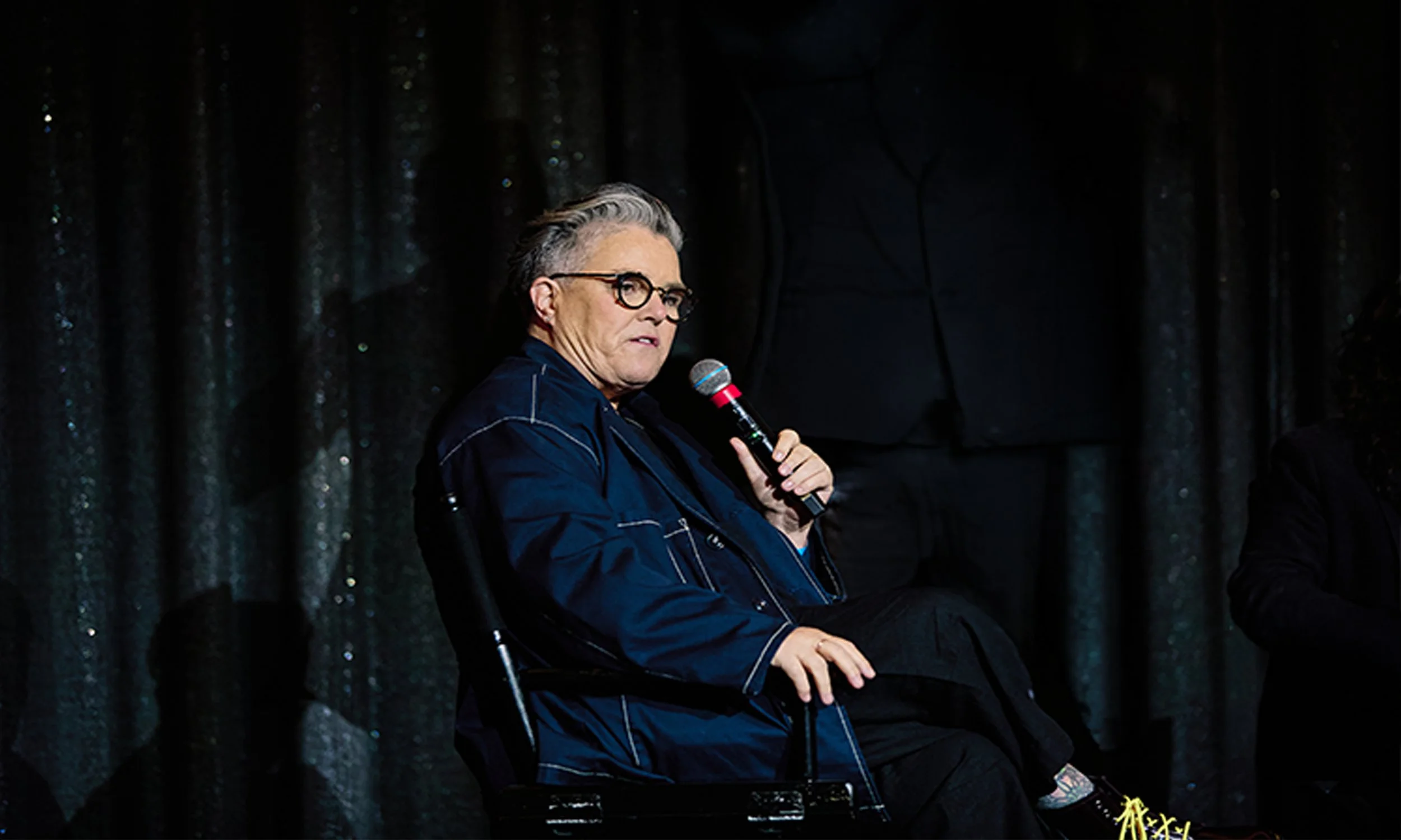 Rosie O'Donnell seated onstage holding a microphone while moderating a Q&A discussion about the award-winning documentary BEING MICHELLE.