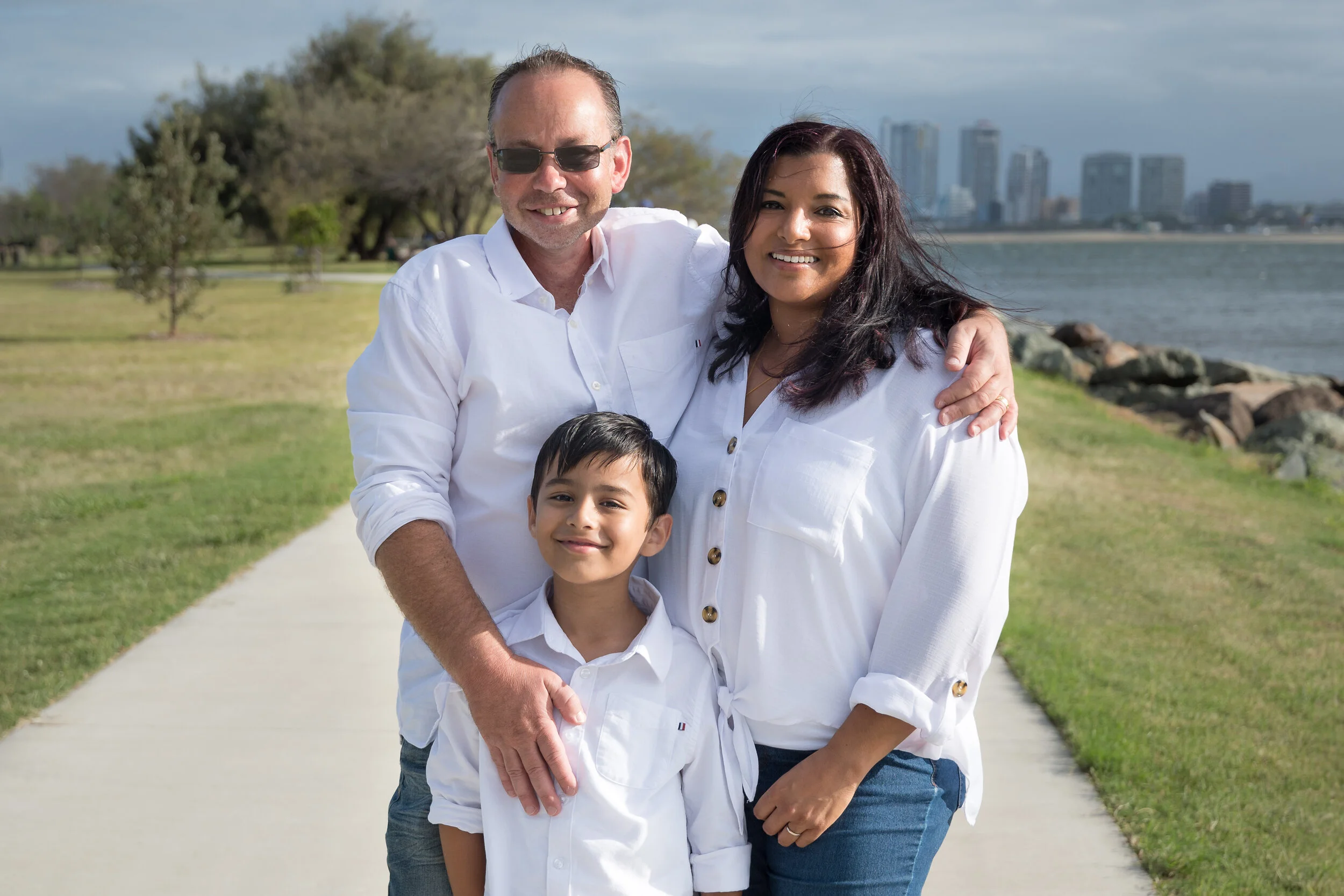 A family of three in white outfits smiling together in a park at Main Beach, with the Gold Coast skyline across the water behind them.