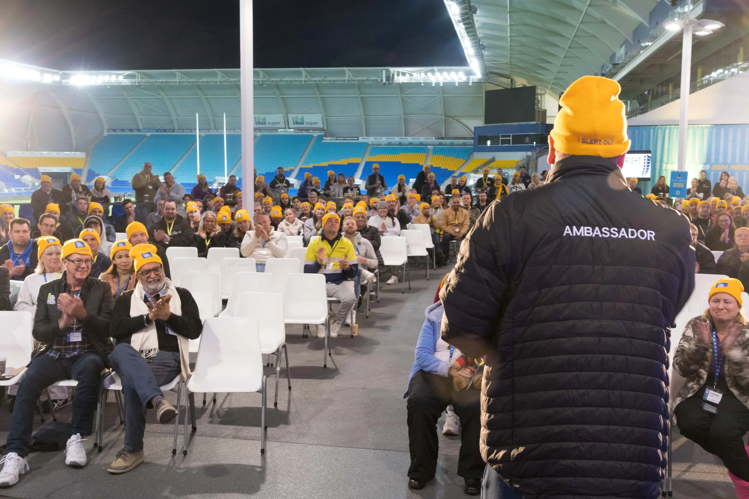 An ambassador presenting to a seated crowd wearing yellow beanies during the CEO Sleepout corporate charity event at Cbus Super Stadium in Robina on the Gold Coast. Captured by event photographer Scott Hardy