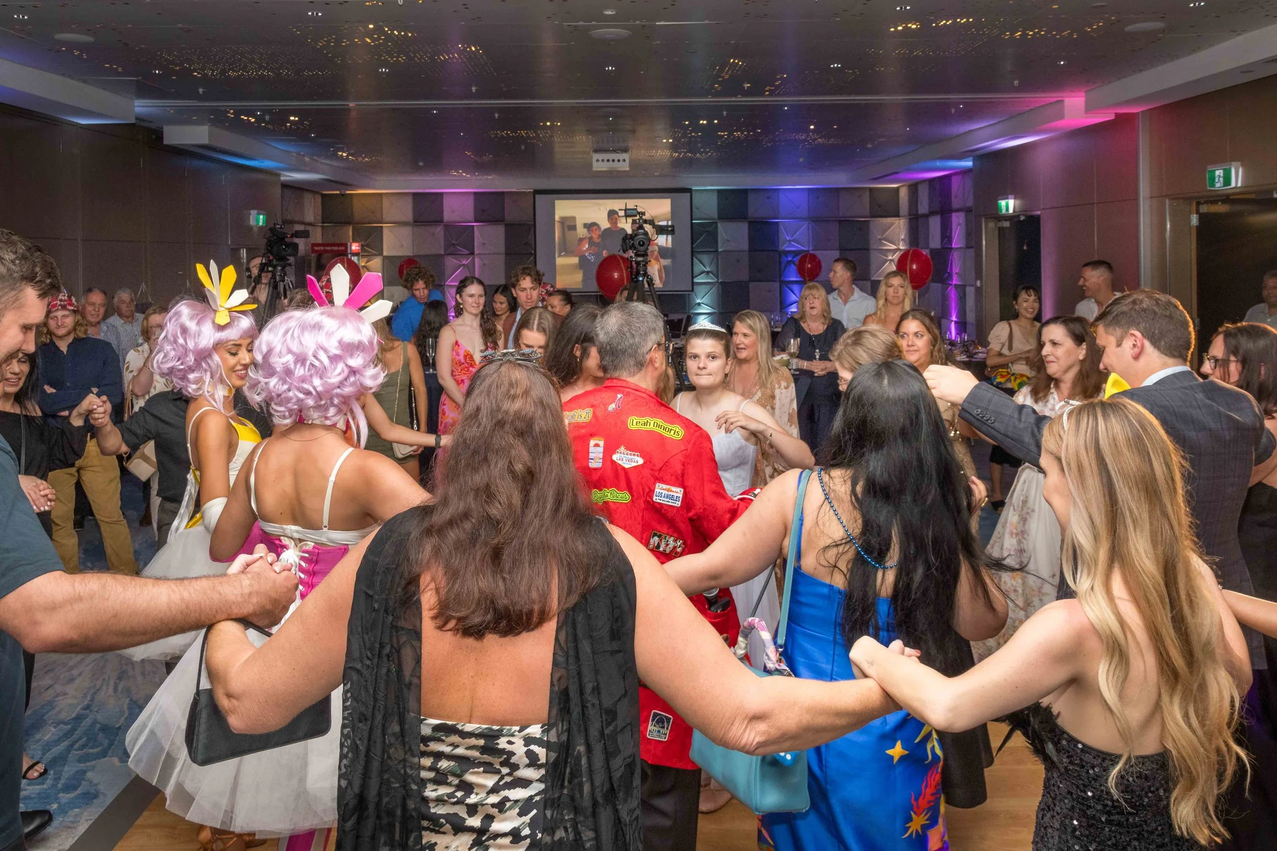 Large group of guests holding hands and dancing in a circle during lively Hilton Ballroom birthday party. 